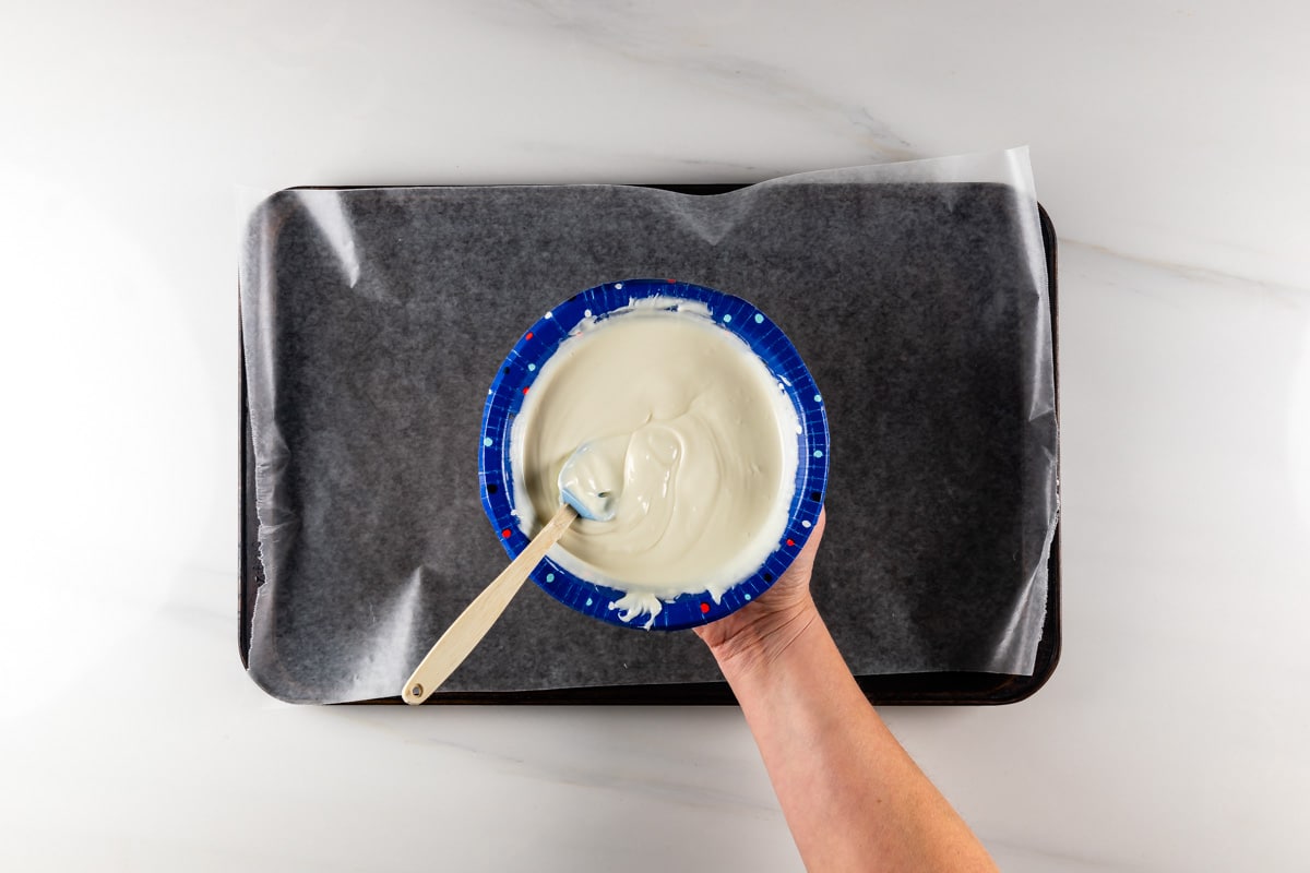 A hand holds a blue bowl filled with white melted chocolate or frosting, with a wooden spoon inside. The bowl is above a baking tray lined with parchment paper on a white marble surface.