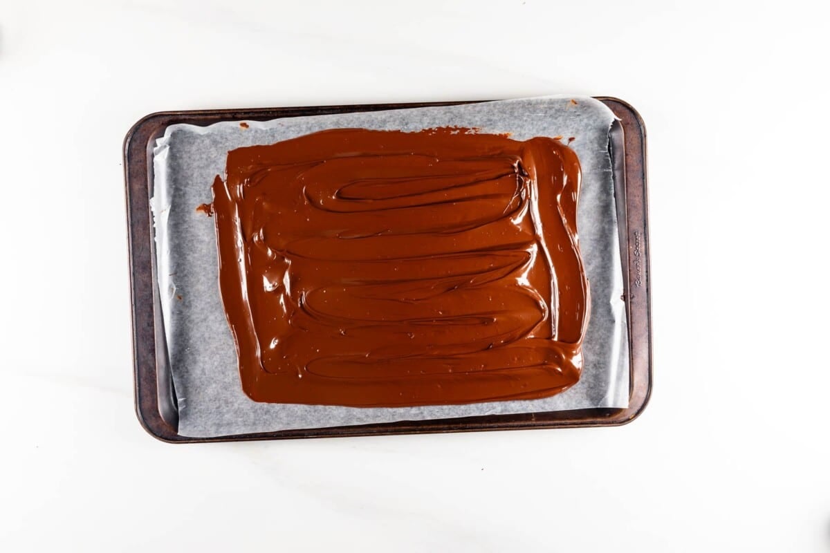 A baking tray lined with parchment paper holds a layer of smooth, melted chocolate spread evenly across its surface.