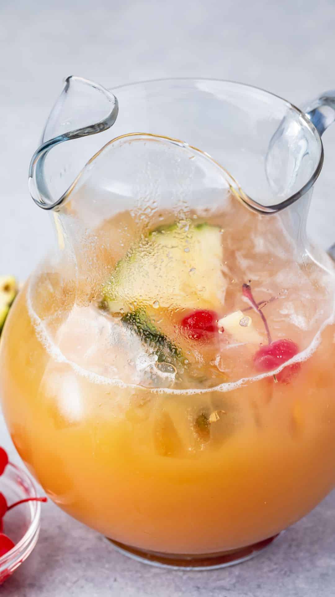 A frosty pitcher filled with an iced fruity punch, featuring slices of pineapple, lemon, and several cherries. The glass pitcher shows condensation, indicating a chilled drink, set against a light background.