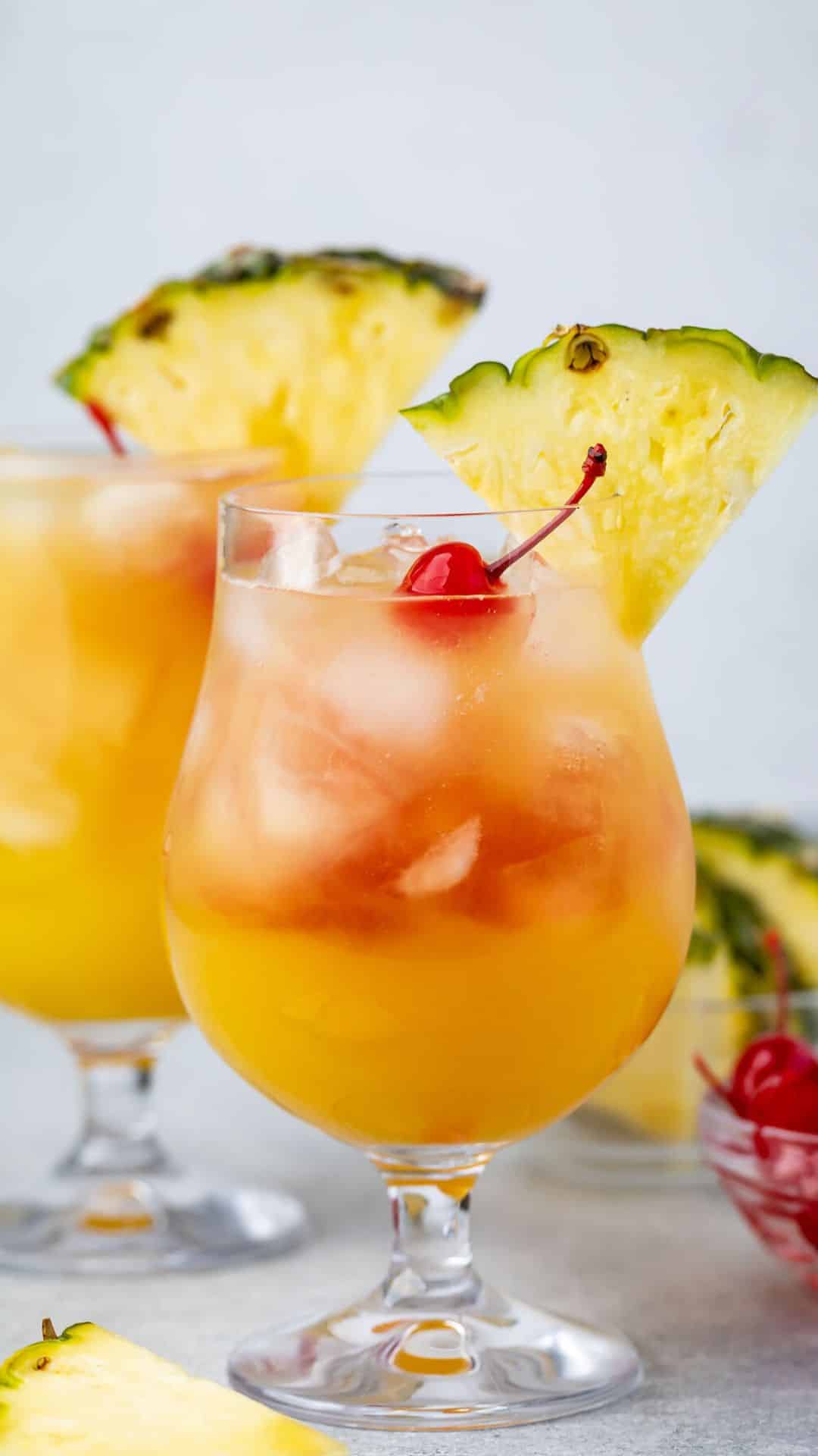 Two glasses of a tropical cocktail garnished with pineapple slices and cherries. The drinks are served over ice, with a vibrant orange hue. A bowl with cherries and pineapple slices is partially visible in the background.