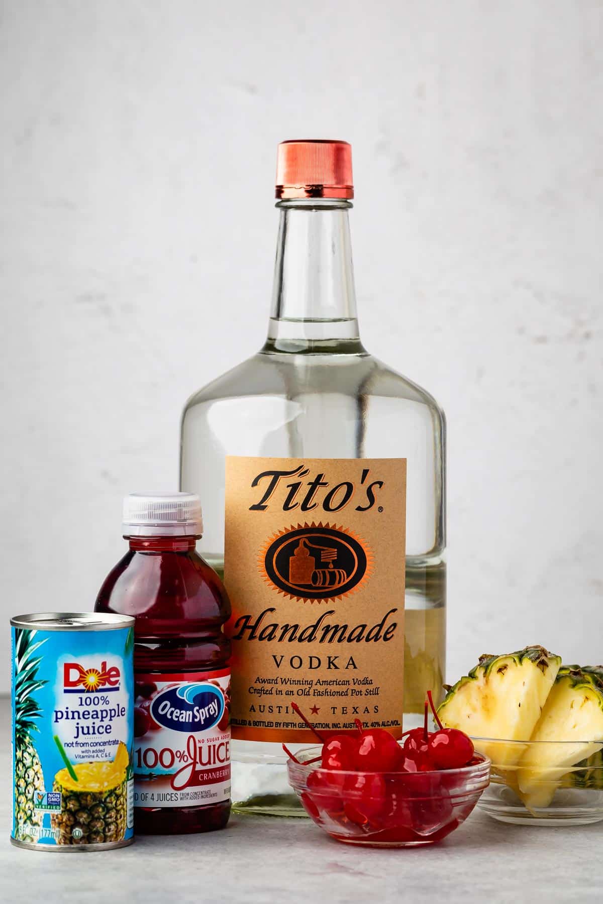 A bottle of Titos Handmade Vodka is surrounded by a can of Dole pineapple juice, a bottle of Ocean Spray cranberry juice, fresh pineapple slices, and a bowl of maraschino cherries on a white background.