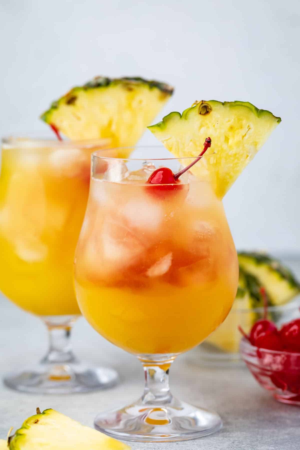 Two glasses of tropical cocktails garnished with pineapple slices and cherries. The drinks are orange with ice cubes, set on a light gray surface. A small bowl of cherries is visible in the background.