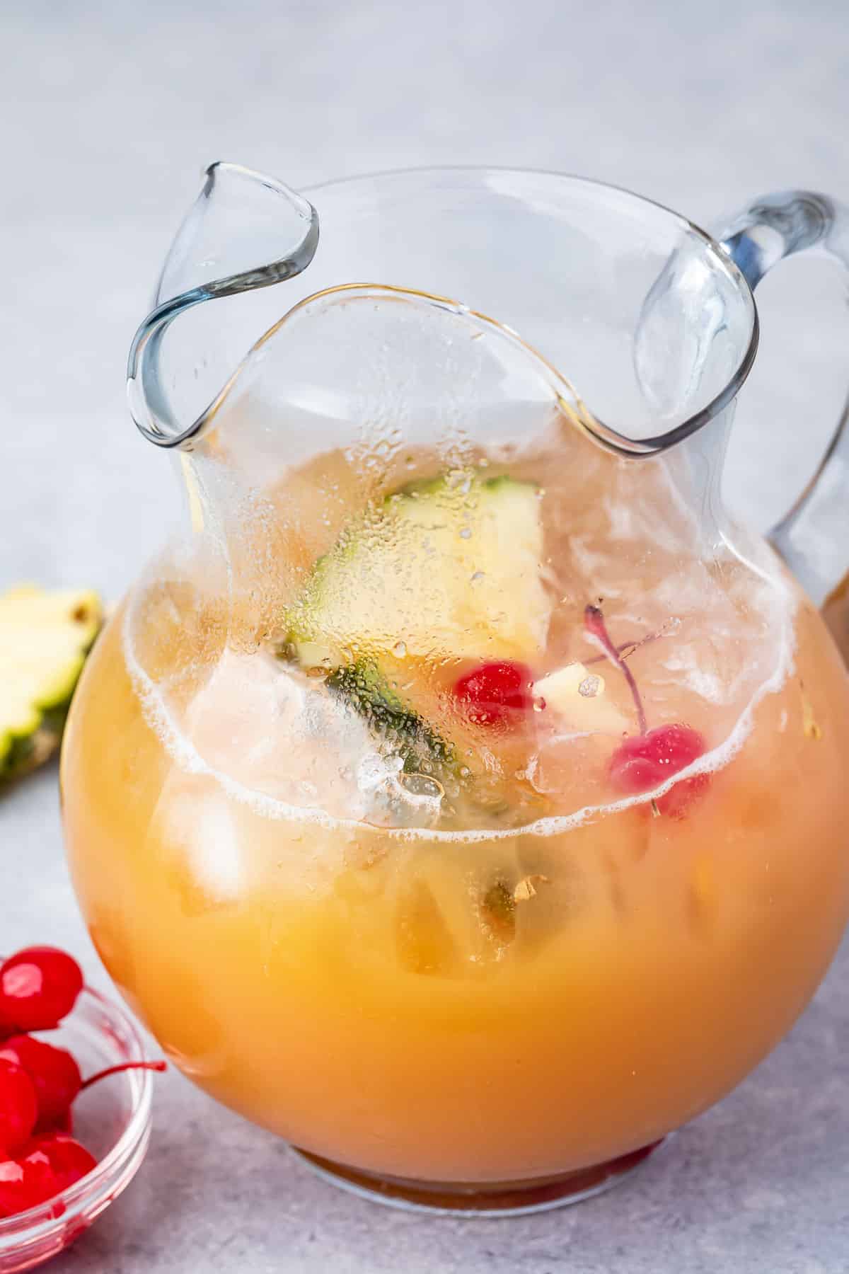 A glass pitcher filled with a tropical fruit punch, featuring pineapple slices, cherries, and ice cubes. A small bowl of red cherries is placed beside the pitcher on a light grey surface.