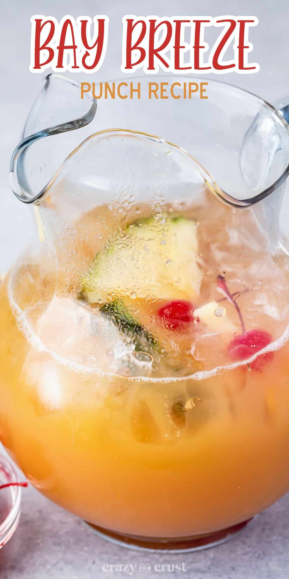 A glass pitcher filled with Bay Breeze Punch, containing pineapple slices, cherries, and ice cubes. The text reads Bay Breeze Punch Recipe at the top. A bright and refreshing drink is visible.