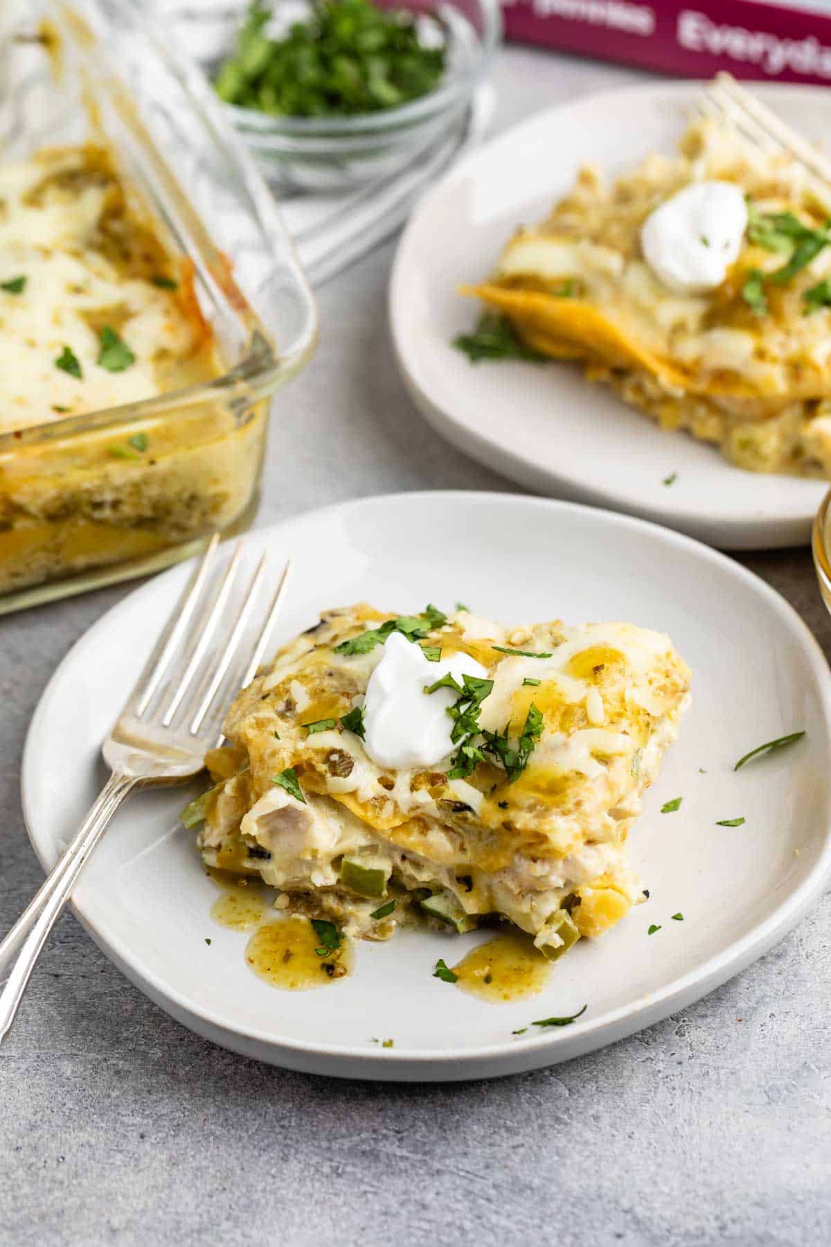 A close-up of two plates each with a serving of creamy chicken enchilada casserole topped with fresh cilantro and dollops of sour cream. A glass baking dish with more casserole is visible in the background. Forks are placed on the plates.