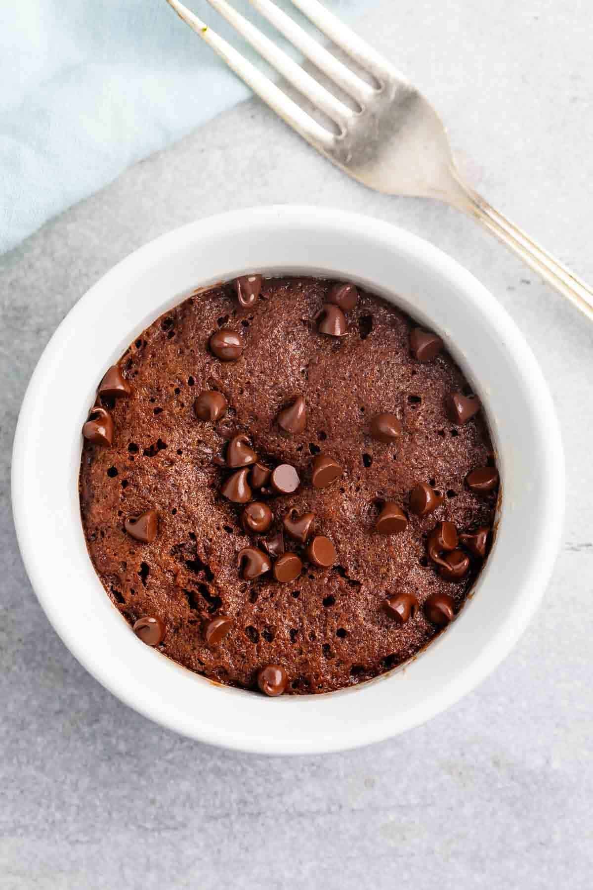A chocolate mug cake topped with chocolate chips in a white mug on a gray stone surface. A fork is placed nearby.