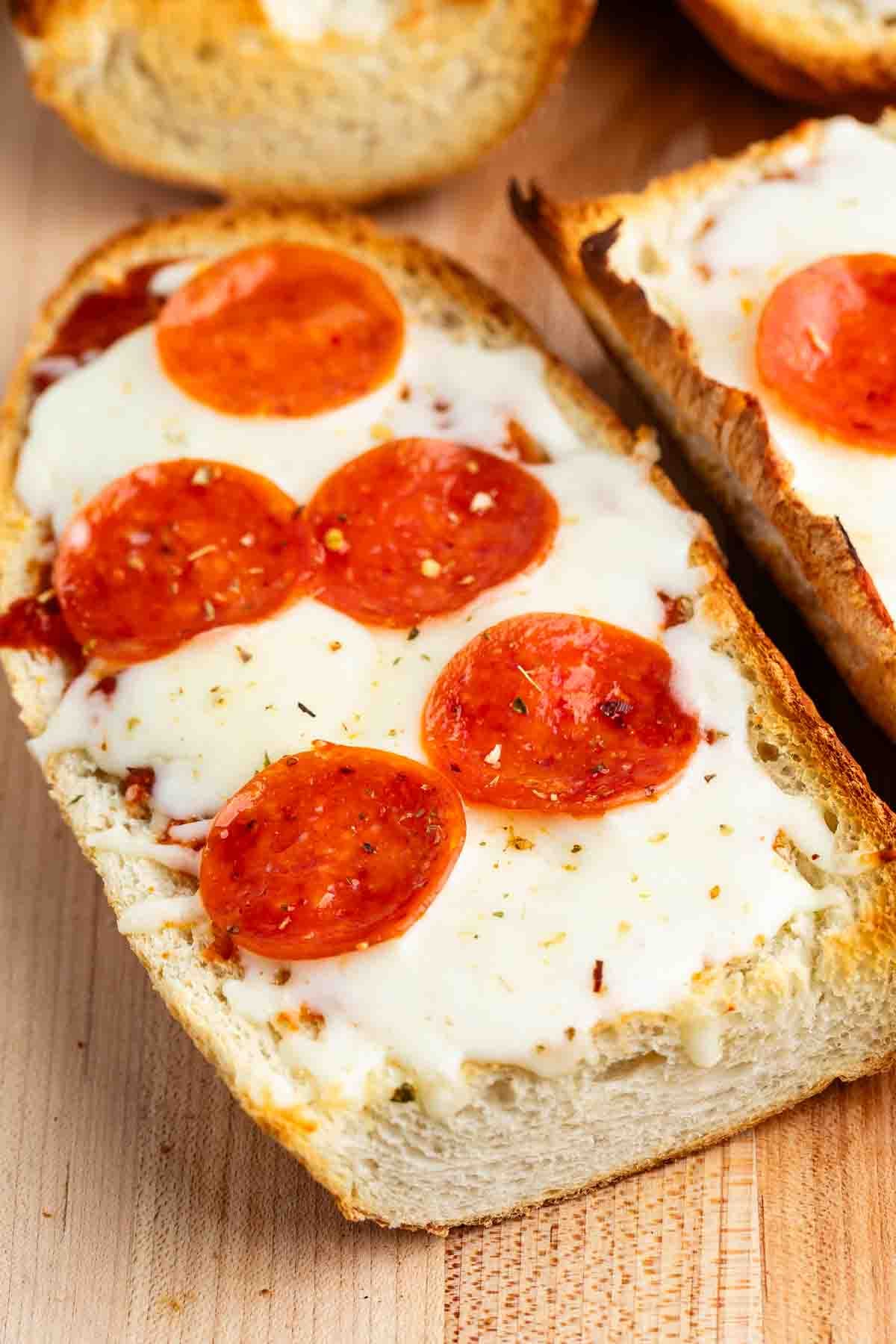 A close-up of French bread pizza topped with melted cheese and pepperoni slices. The bread is toasted, and the pizza is sprinkled with herbs. It rests on a wooden surface.