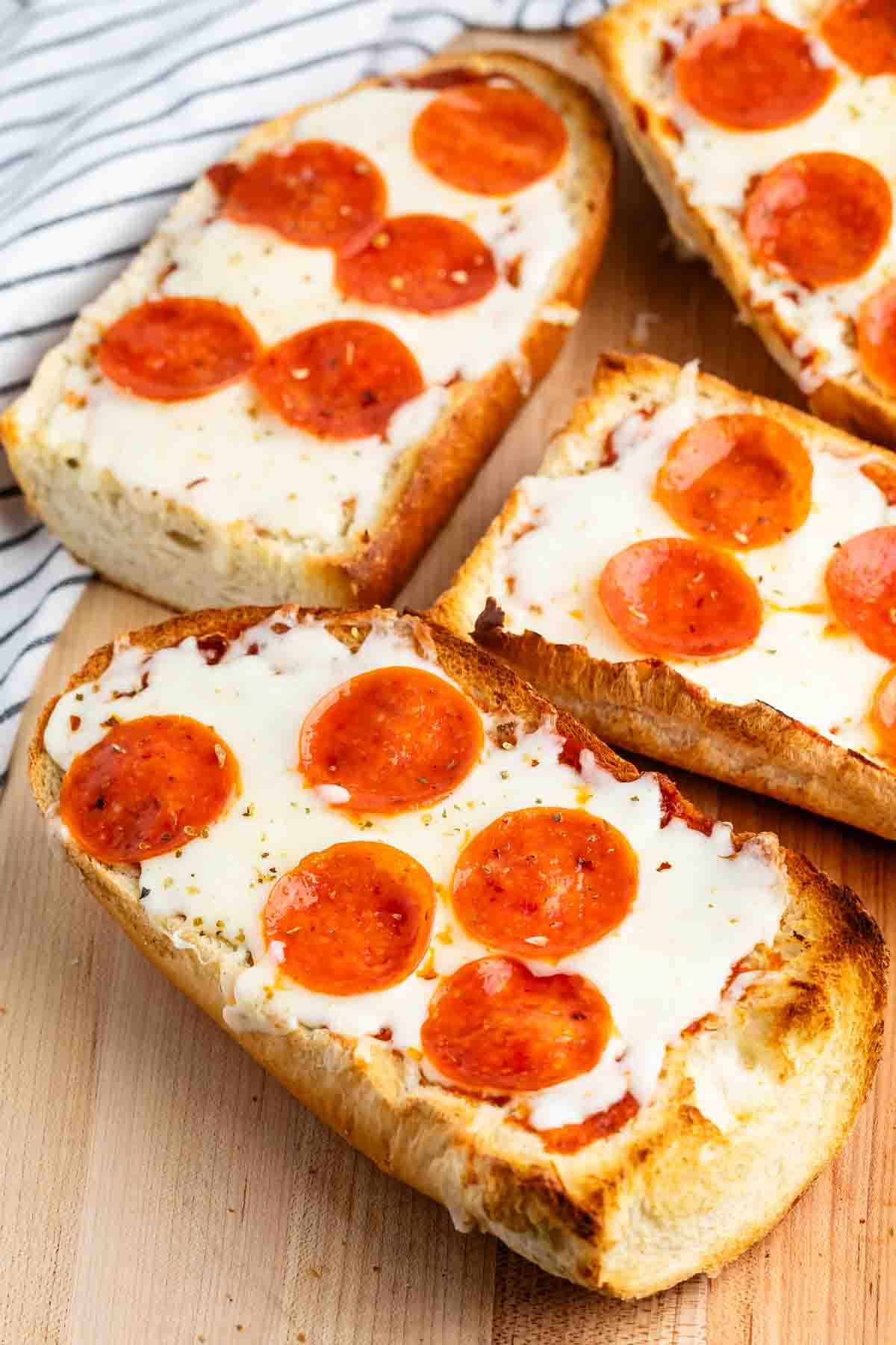 Slices of bread topped with melted cheese and pepperoni are arranged on a wooden surface. A striped cloth is partially visible in the background.