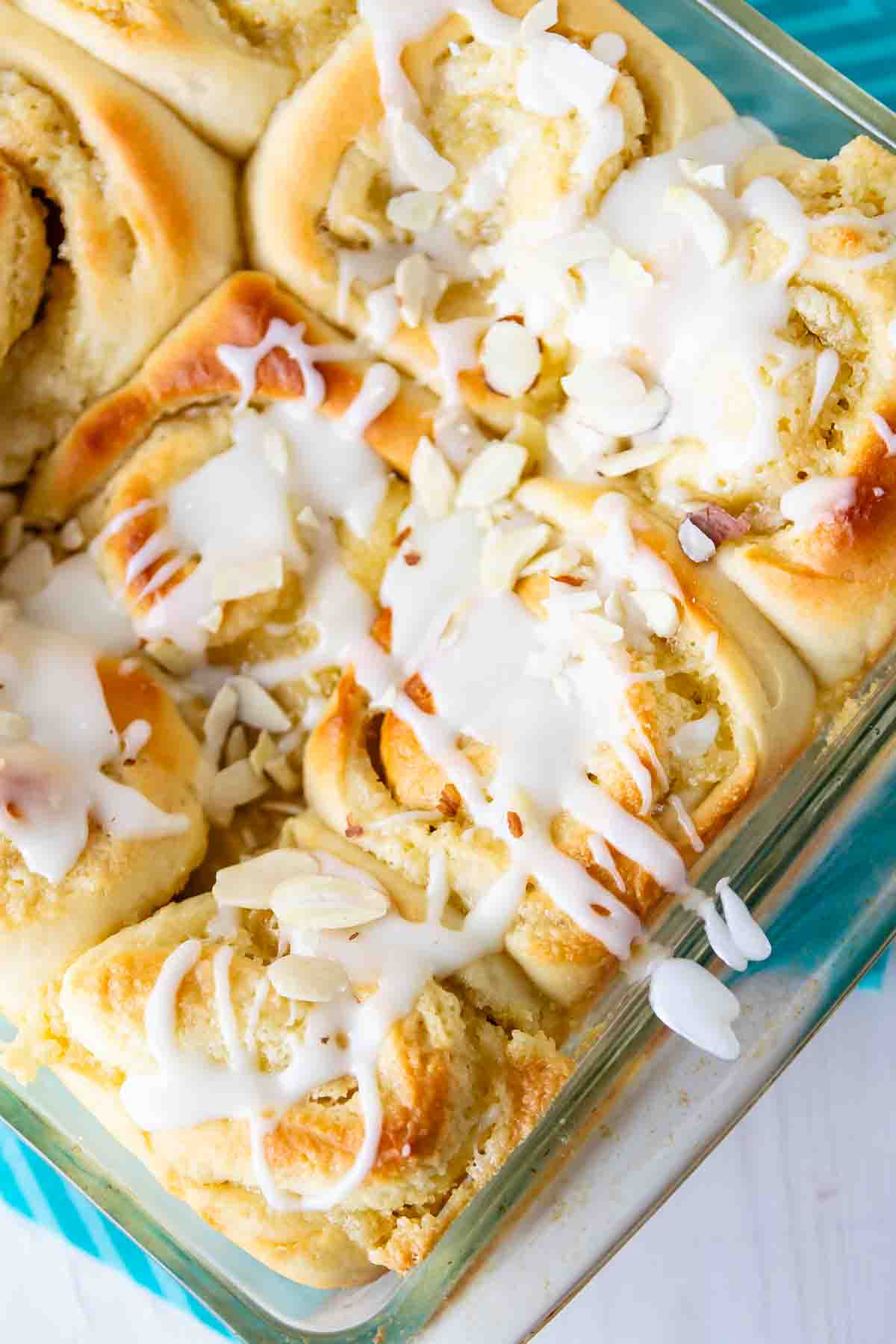 All the flavors of an almond croissant...in a sweet roll! Close-up of freshly baked cinnamon rolls in a glass dish, topped with white icing and sliced almonds. The rolls are golden brown, with a soft and fluffy texture visible under the glaze.