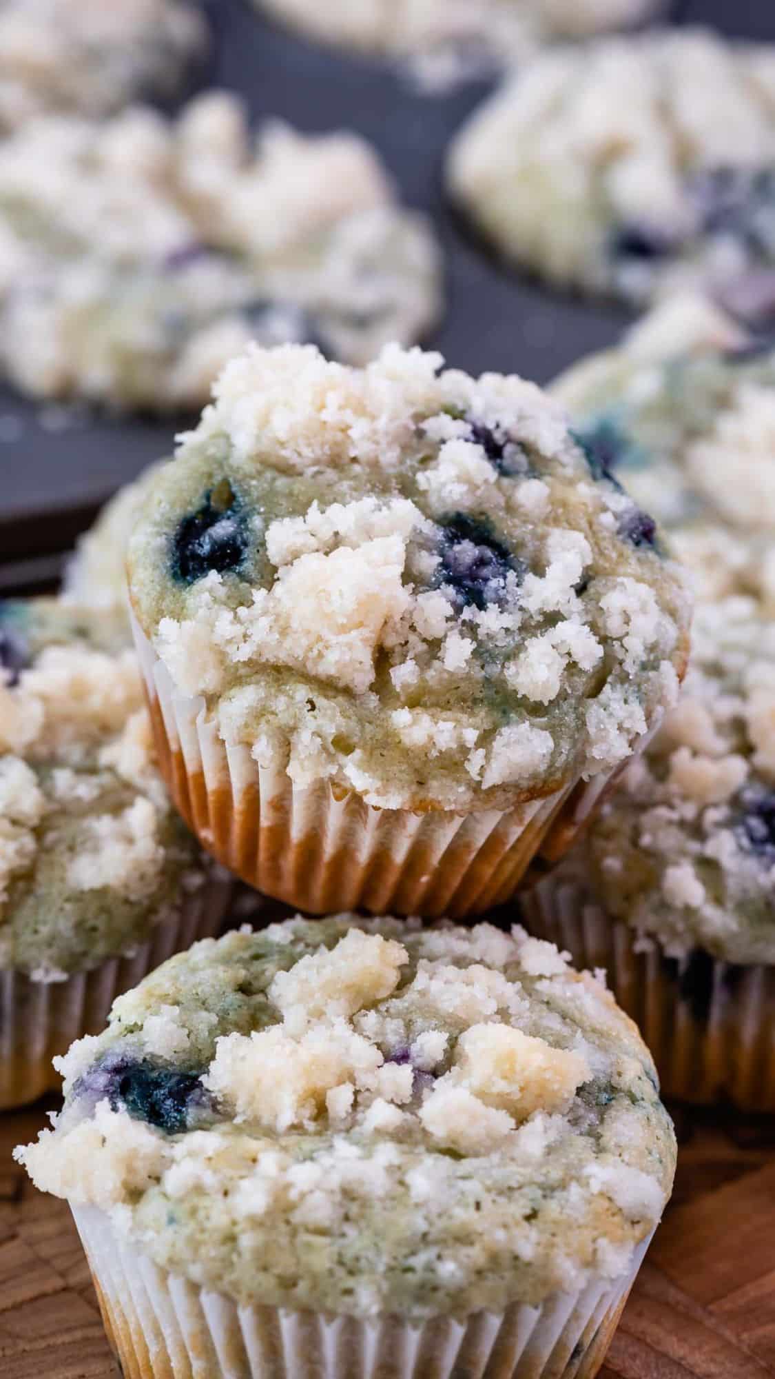 Three blueberry muffins topped with crumbly streusel, arranged in a stack. More muffins are visible in the background on a baking tray.