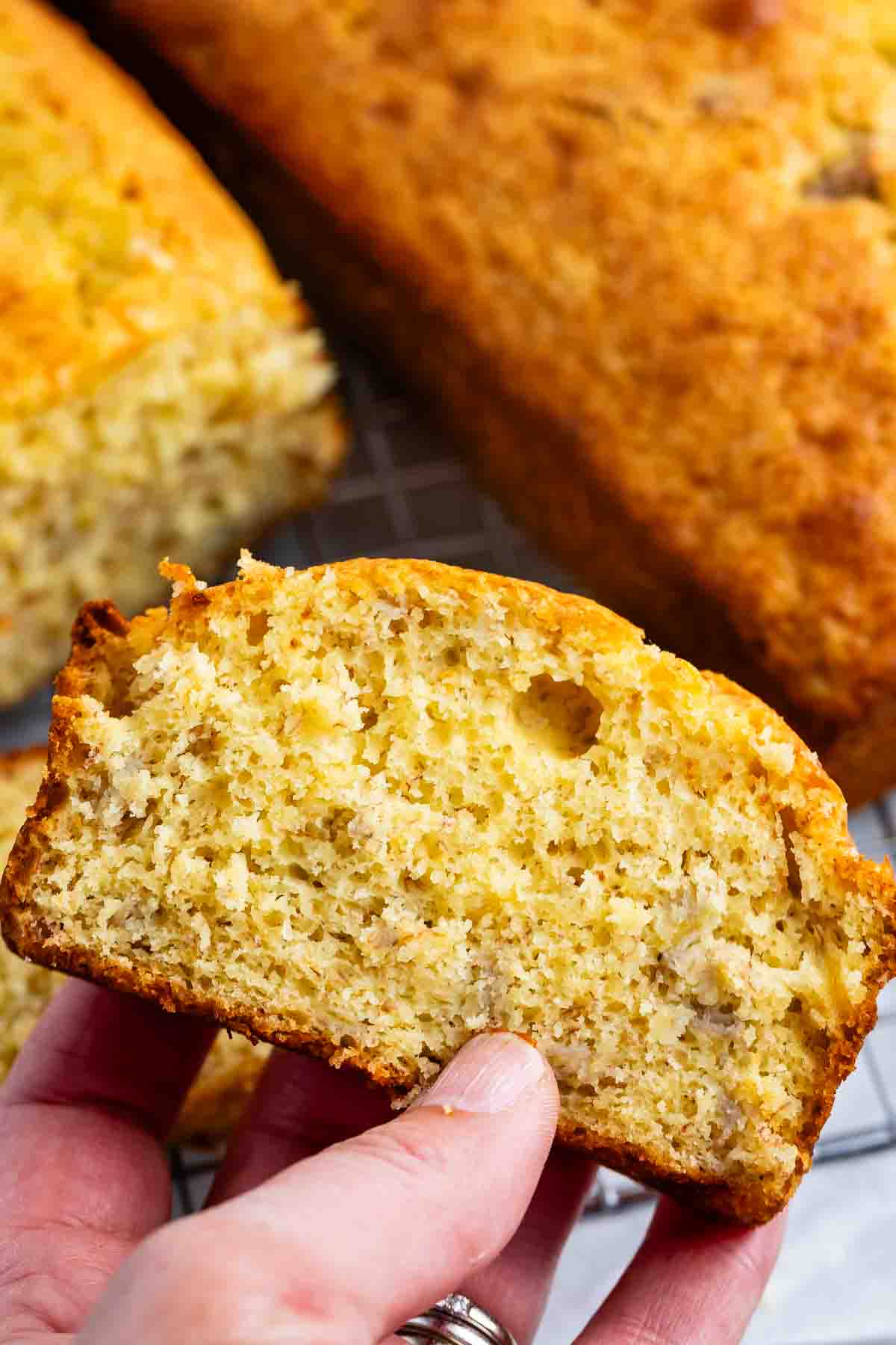 A hand holding a slice of banana bread in the foreground, showing its moist texture with visible banana pieces. In the background, more banana bread is placed on a cooling rack.