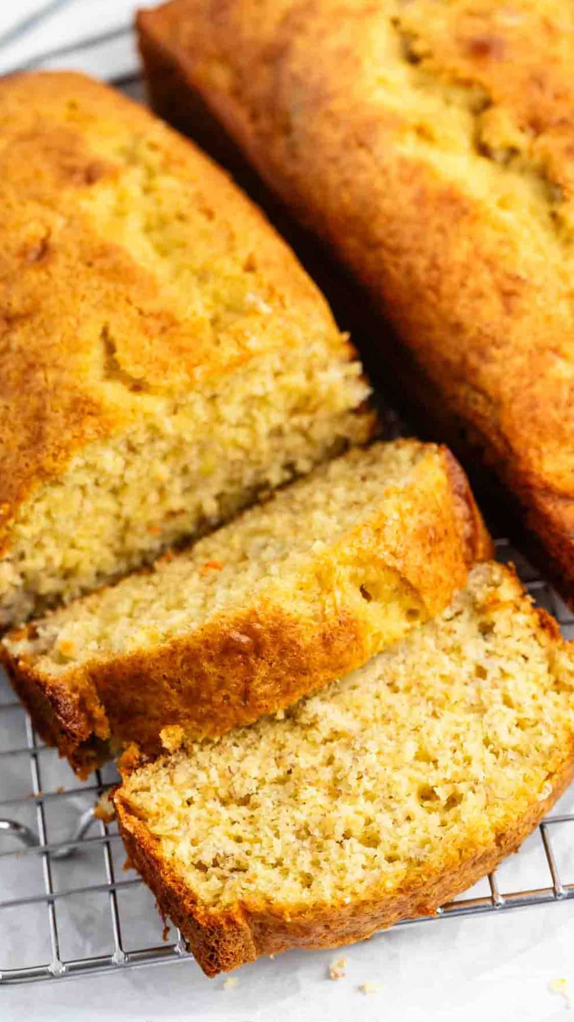 Three freshly baked banana bread loaves on a cooling rack, with one loaf partially sliced, revealing a moist and textured interior. The golden-brown crust looks crisp and inviting.