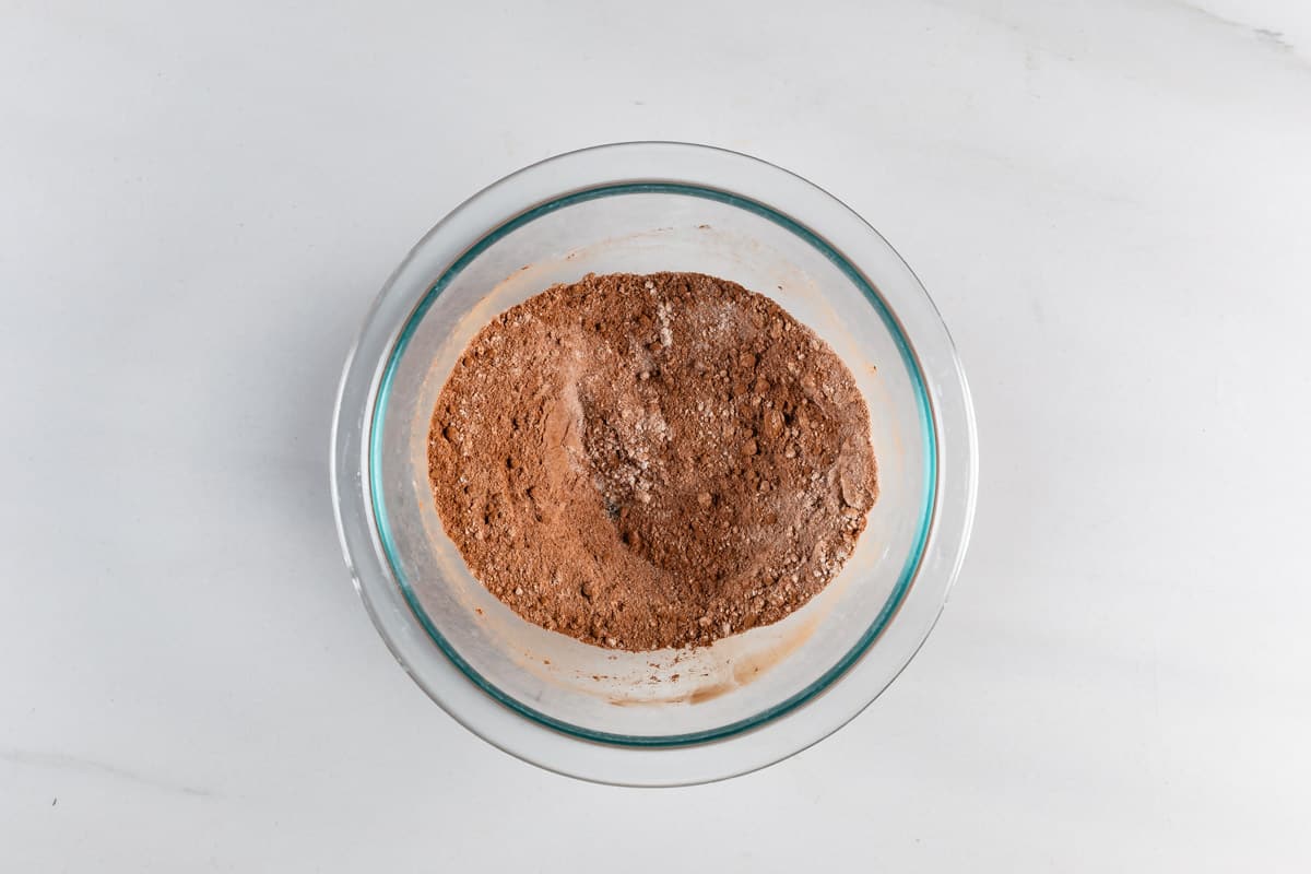 A glass bowl containing a mixture of dry ingredients, primarily cocoa powder, sits on a white surface. The powdery texture and uniform color suggest preparation for baking.