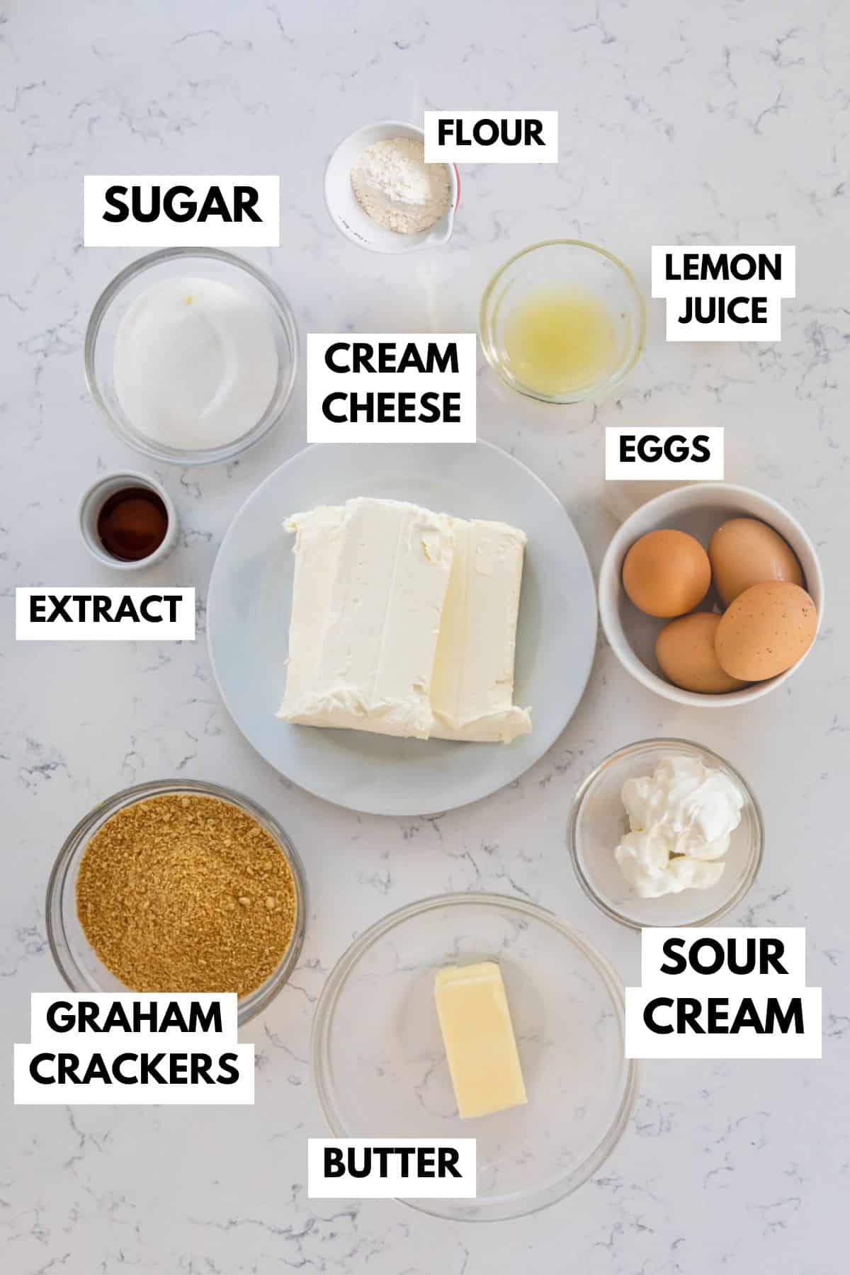 A collection of ingredients for the best cheesecake recipe sits on a marble countertop: sugar, flour, lemon juice, cream cheese blocks, eggs, sour cream, butter, graham crackers, and vanilla extract, each neatly labeled in a small dish.