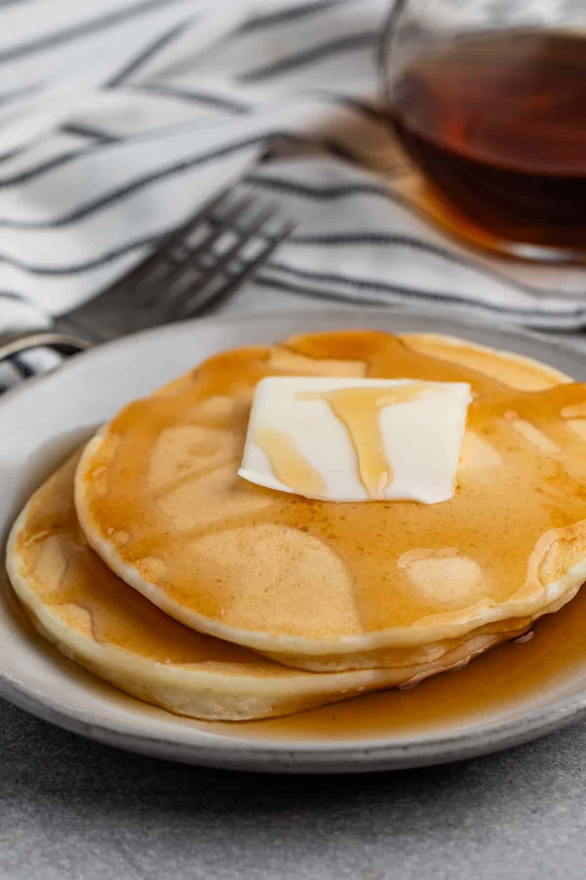 A stack of pancakes topped with a pat of butter and drizzled with syrup on a white plate. A fork and a glass container of syrup are in the background, placed on a striped cloth.