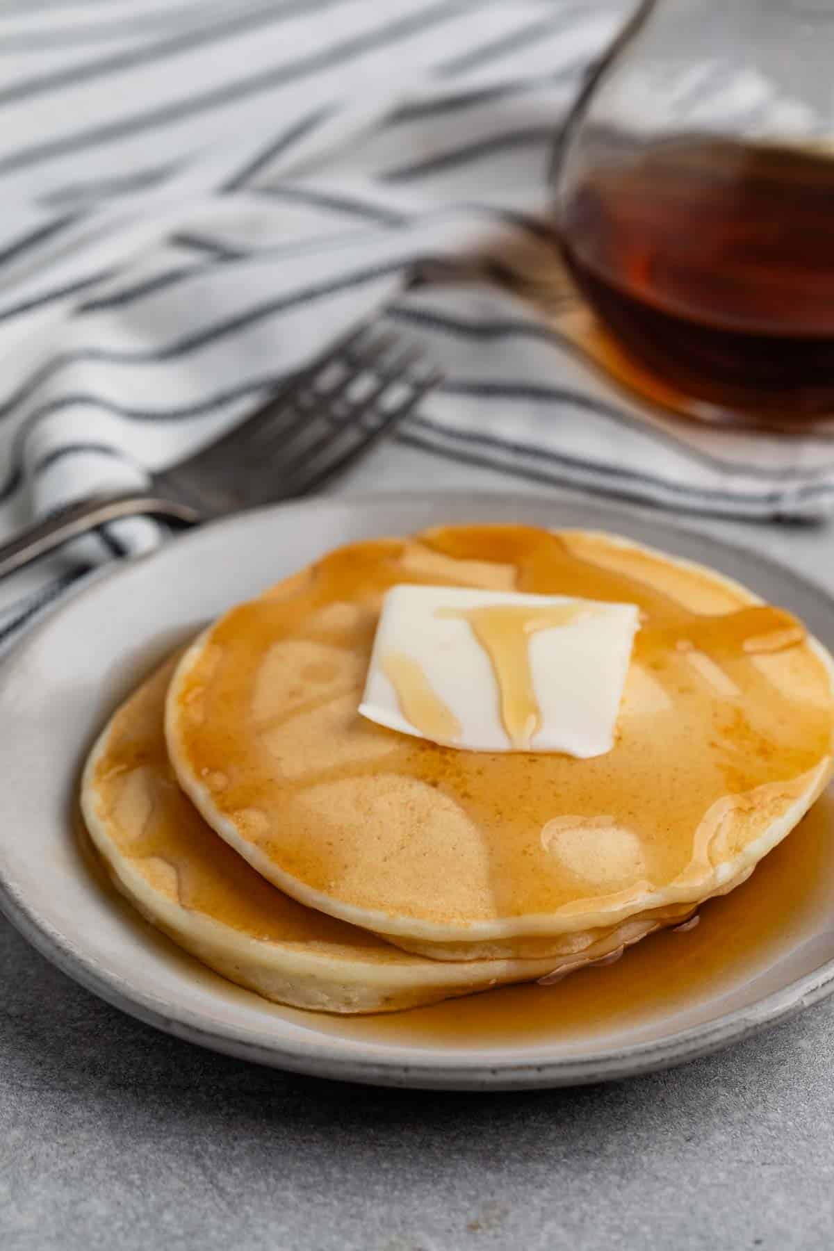 A stack of pancakes topped with a pat of butter and drizzled with syrup on a white plate. A glass syrup container and a striped napkin with a fork are in the background.