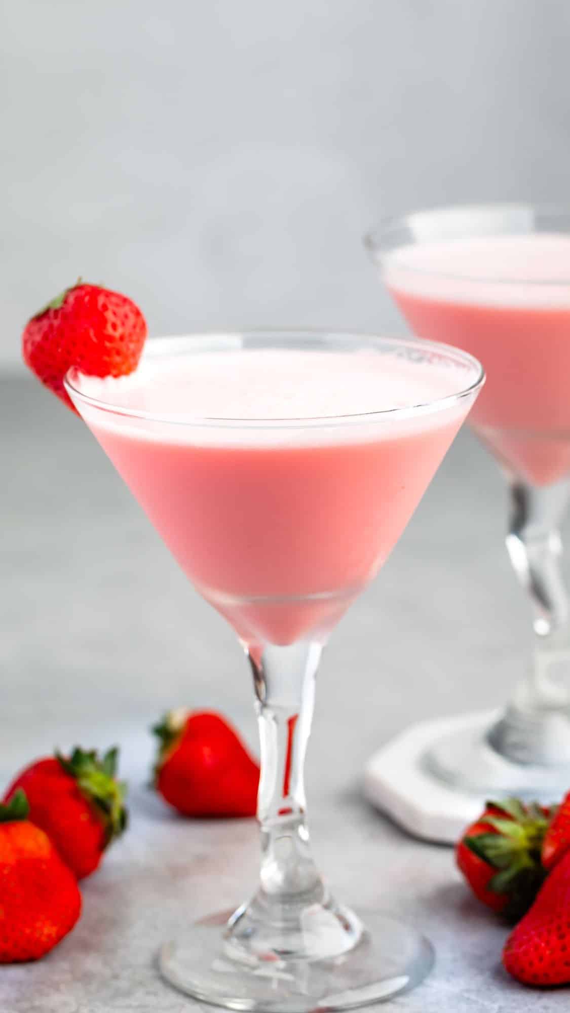 Two pink cocktails in martini glasses, each topped with a strawberry on the rim. More strawberries are scattered on the gray surface. The background is blurred, highlighting the drinks.