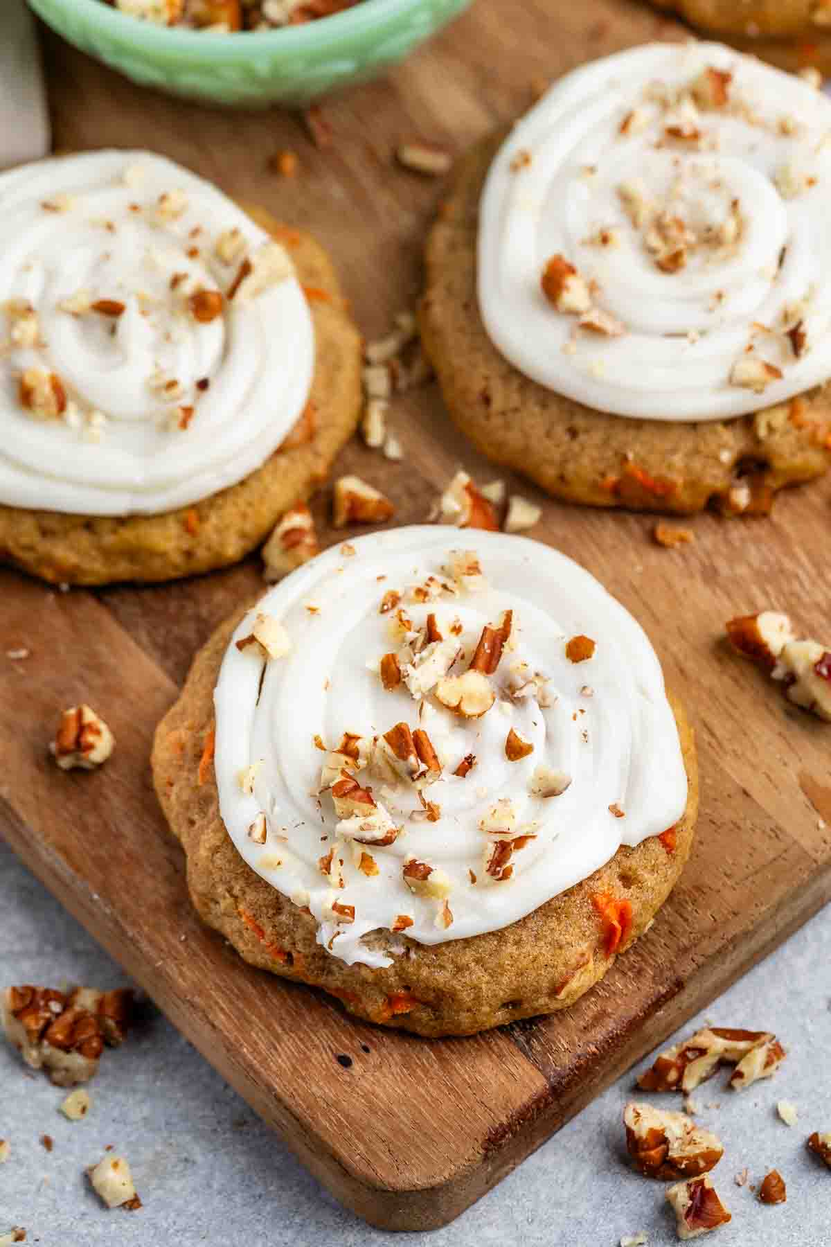 Carrot cake cookies with white frosting swirl and chopped nuts are displayed on a wooden board. A green bowl with more nuts is in the background. The cookies have visible carrot pieces and a soft, rustic appearance.