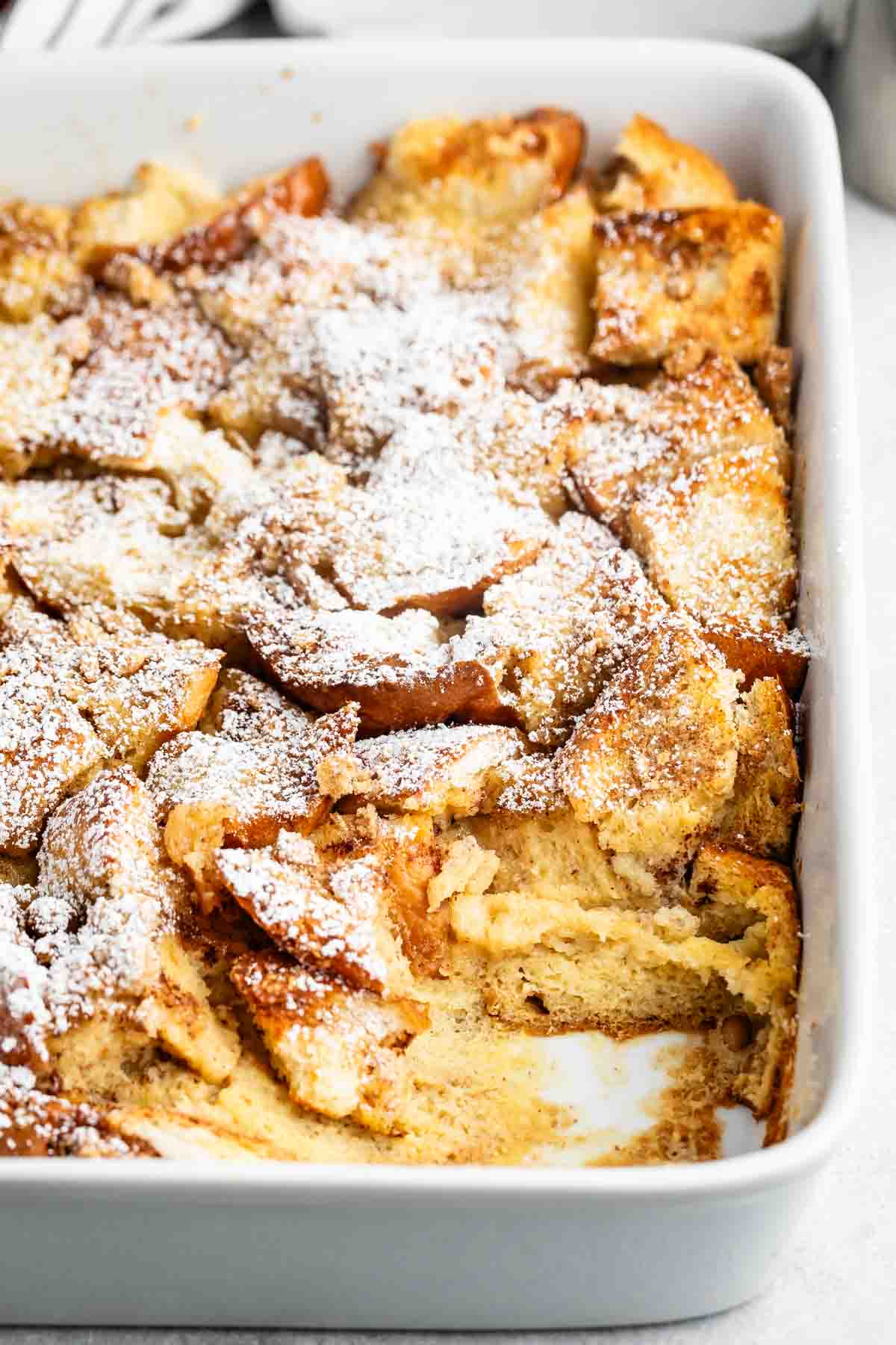 A baked bread pudding in a rectangular white dish, topped with a dusting of powdered sugar. Some pieces are golden-brown and slightly crispy. A section is missing, revealing the soft, custardy interior.