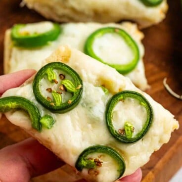 A hand holding a slice of bread topped with melted cheese and sliced jalapeños. Additional slices are visible in the background on a wooden surface.