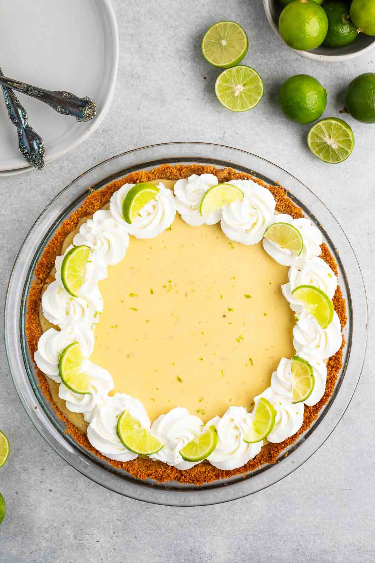 A Key lime pie garnished with whipped cream and lime slices sits on a gray surface. Beside the pie are whole and halved limes. Two decorative spoons rest on a stack of white plates in the background.