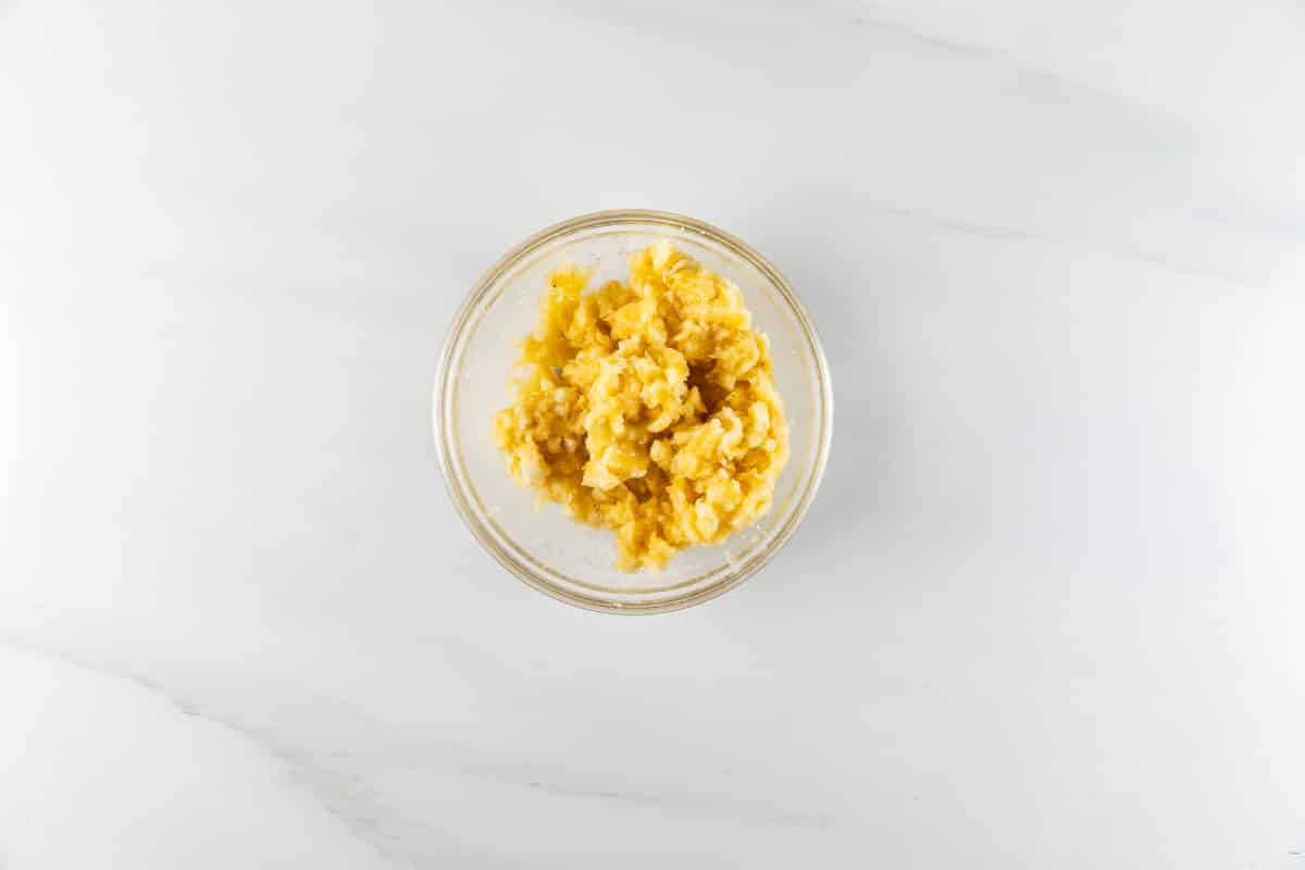A small glass bowl filled with creamy, golden-yellow scrambled eggs sits on a smooth, light gray marble surface. The eggs appear fluffy and slightly textured, and the setting is minimalistic and clean.