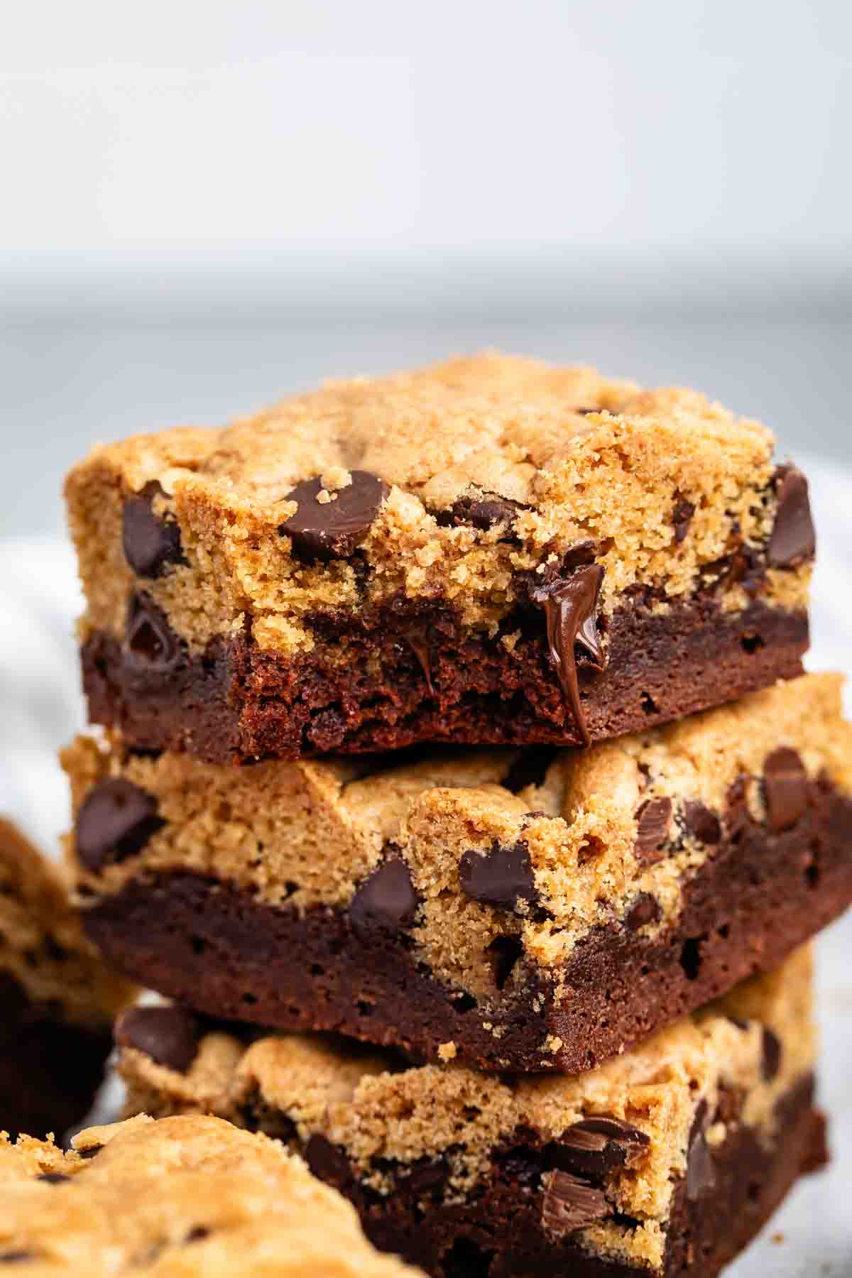 Stacked squares of brookies, a dessert combining chocolate chip cookies and brownies, are shown. The top layer is cookie dough with chocolate chips, while the bottom layer is brownie.