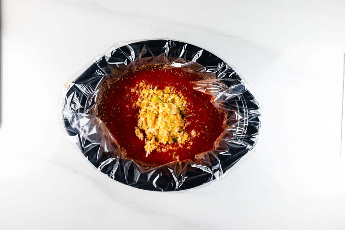 A slow cooker lined with a plastic liner contains a mixture of red sauce and chopped vegetables. The ingredients are set against a white countertop, ready for cooking.