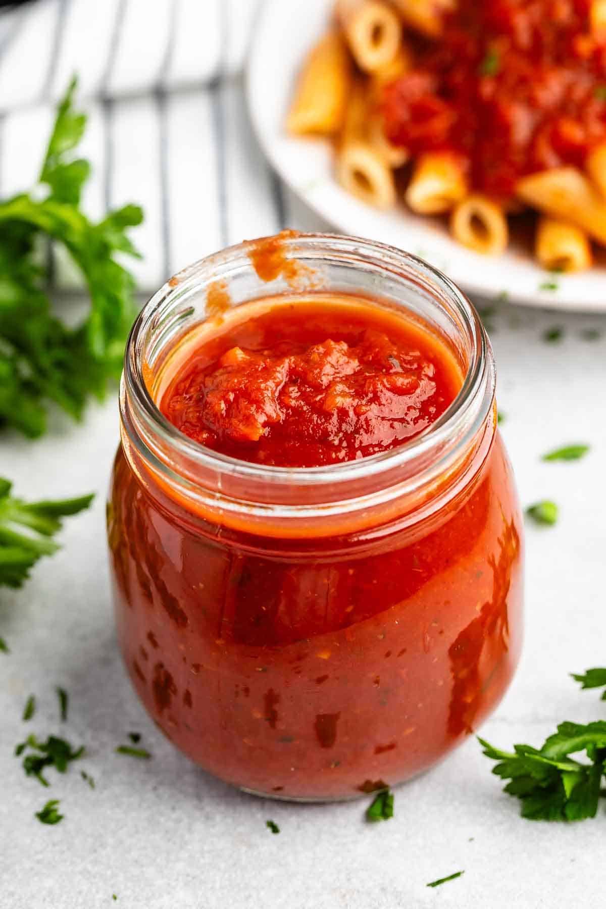 A jar of red marinara sauce sits on a white surface surrounded by sprigs of parsley. In the background, a plate of pasta topped with the same sauce is visible, partially blurred.