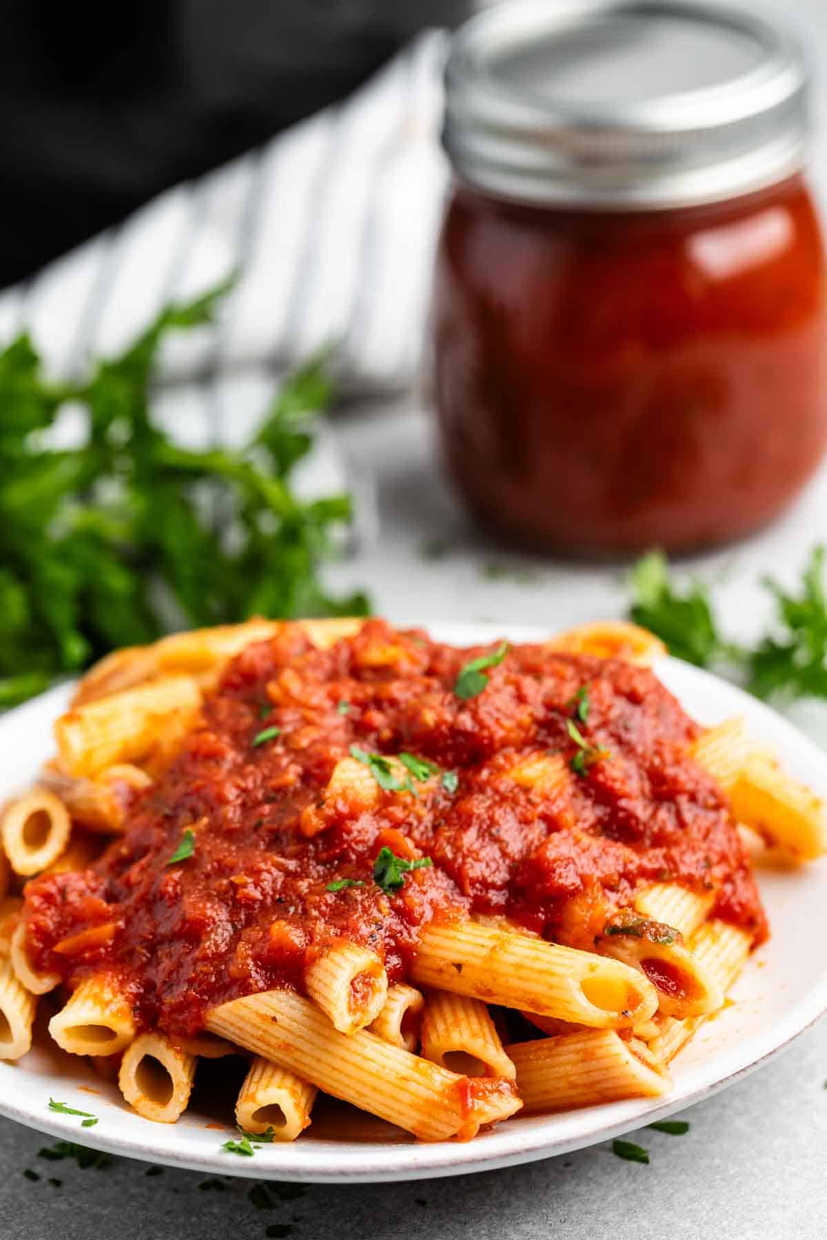 A plate of penne pasta topped with rich tomato sauce, garnished with herbs. In the background, a jar of sauce and fresh herbs are visible.