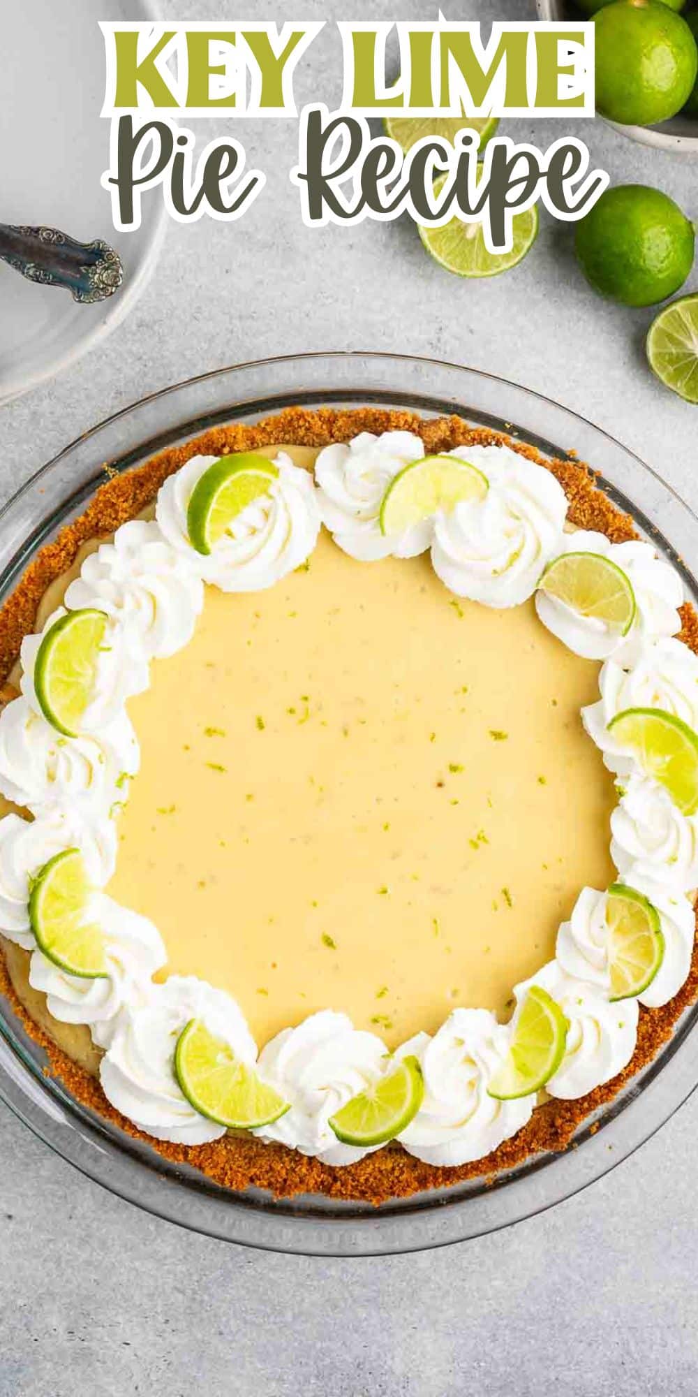 Key lime pie with a creamy filling, topped with whipped cream and slices of lime, in a graham cracker crust. The text Key Lime Pie Recipe is at the top. A few whole limes and a pie server are on the gray surface nearby.
