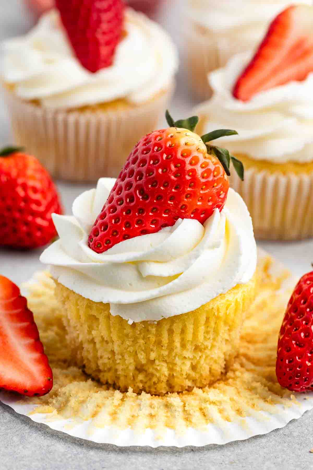 Vanilla cupcake with white frosting topped with a fresh strawberry, surrounded by more cupcakes.