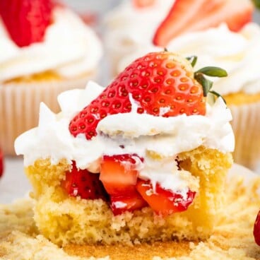 Vanilla cupcake with white frosting topped with a fresh strawberry, surrounded by more cupcakes.