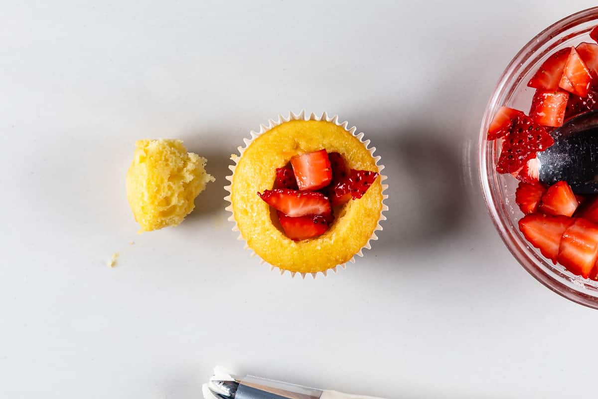 vanilla cupcake with center removed from it and filled with chopped strawberries. Next to it is a bowl of chopped strawberries.