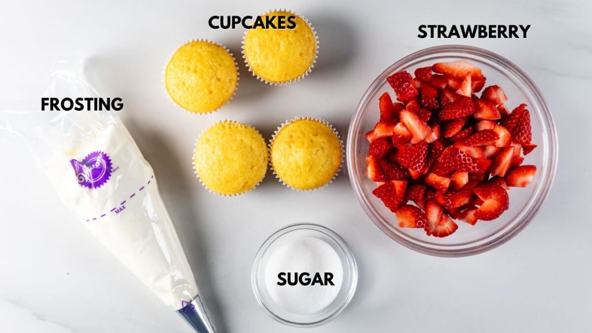 Top view of baking ingredients on a white surface: three plain cupcakes, a bowl of sliced strawberries, a bowl of sugar, and a piping bag filled with frosting. Each item is labeled.