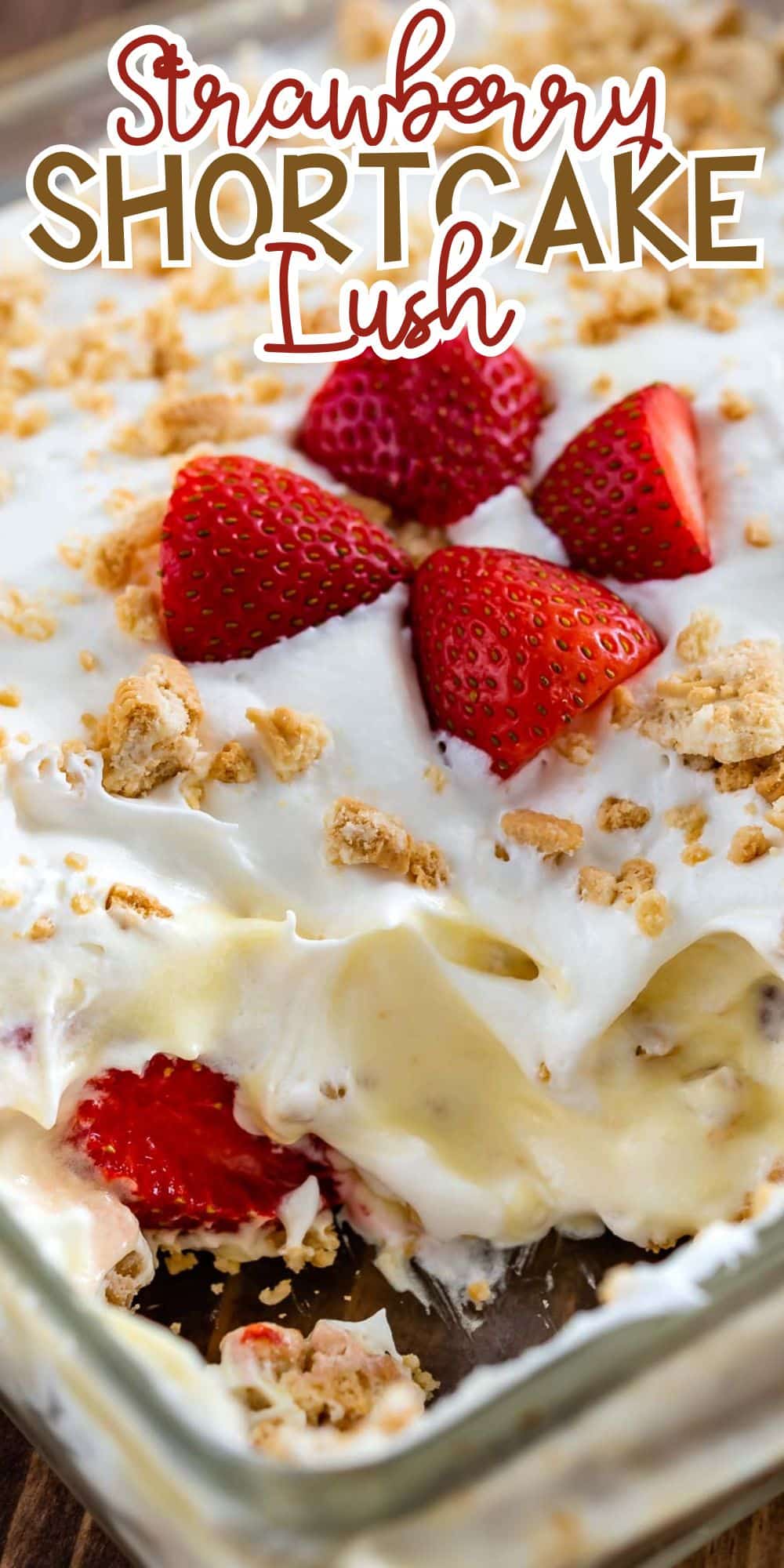 A glass dish of strawberry shortcake lush dessert, featuring layers of whipped cream, pudding, and strawberry slices. Crumbled cookies are sprinkled on top, and the dish has a corner portion missing, revealing its layered texture.