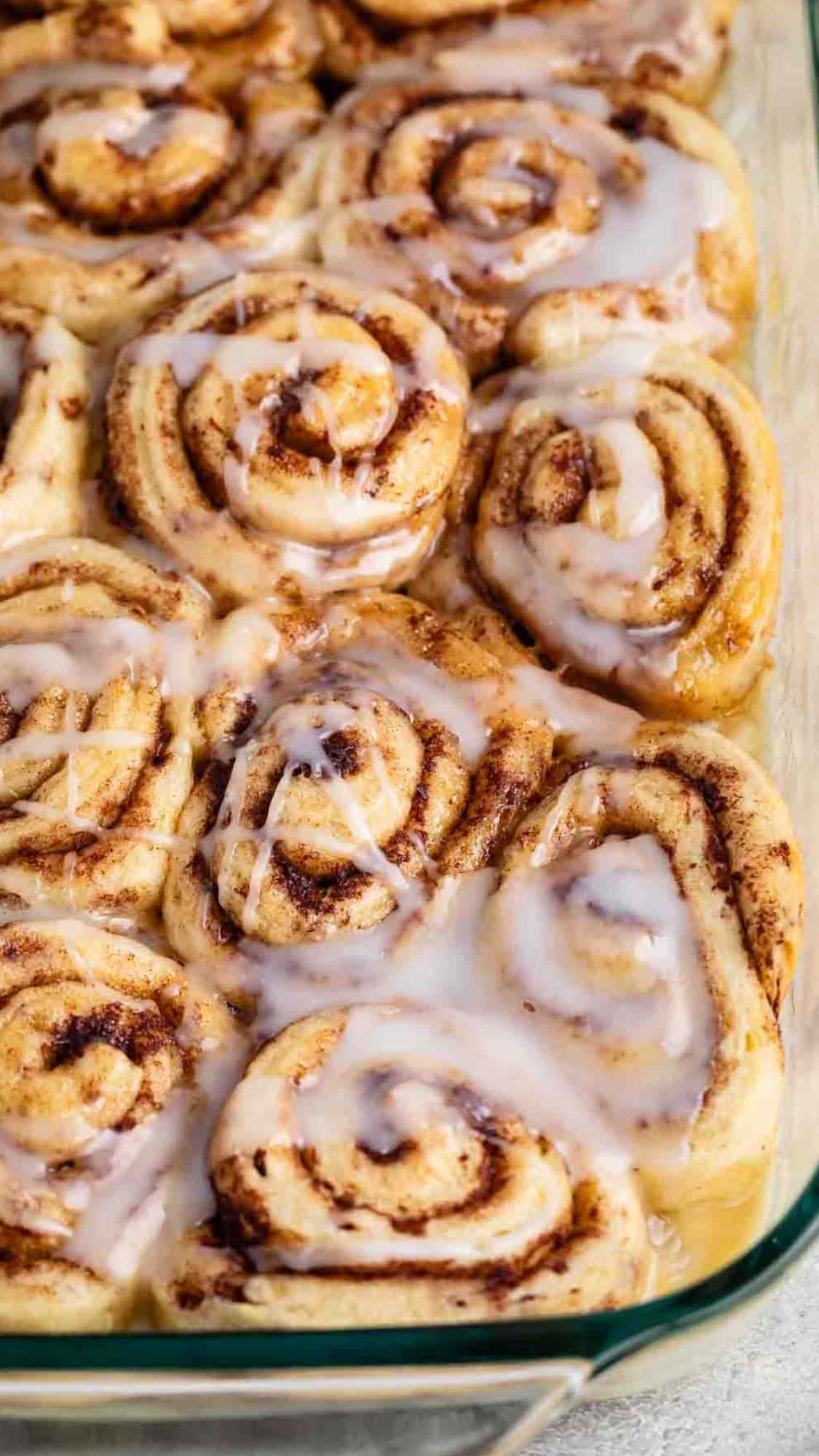 A close-up of freshly baked cinnamon rolls in a glass baking dish. The rolls are swirled with cinnamon and topped with a glossy layer of melting icing, giving them a warm and inviting appearance.