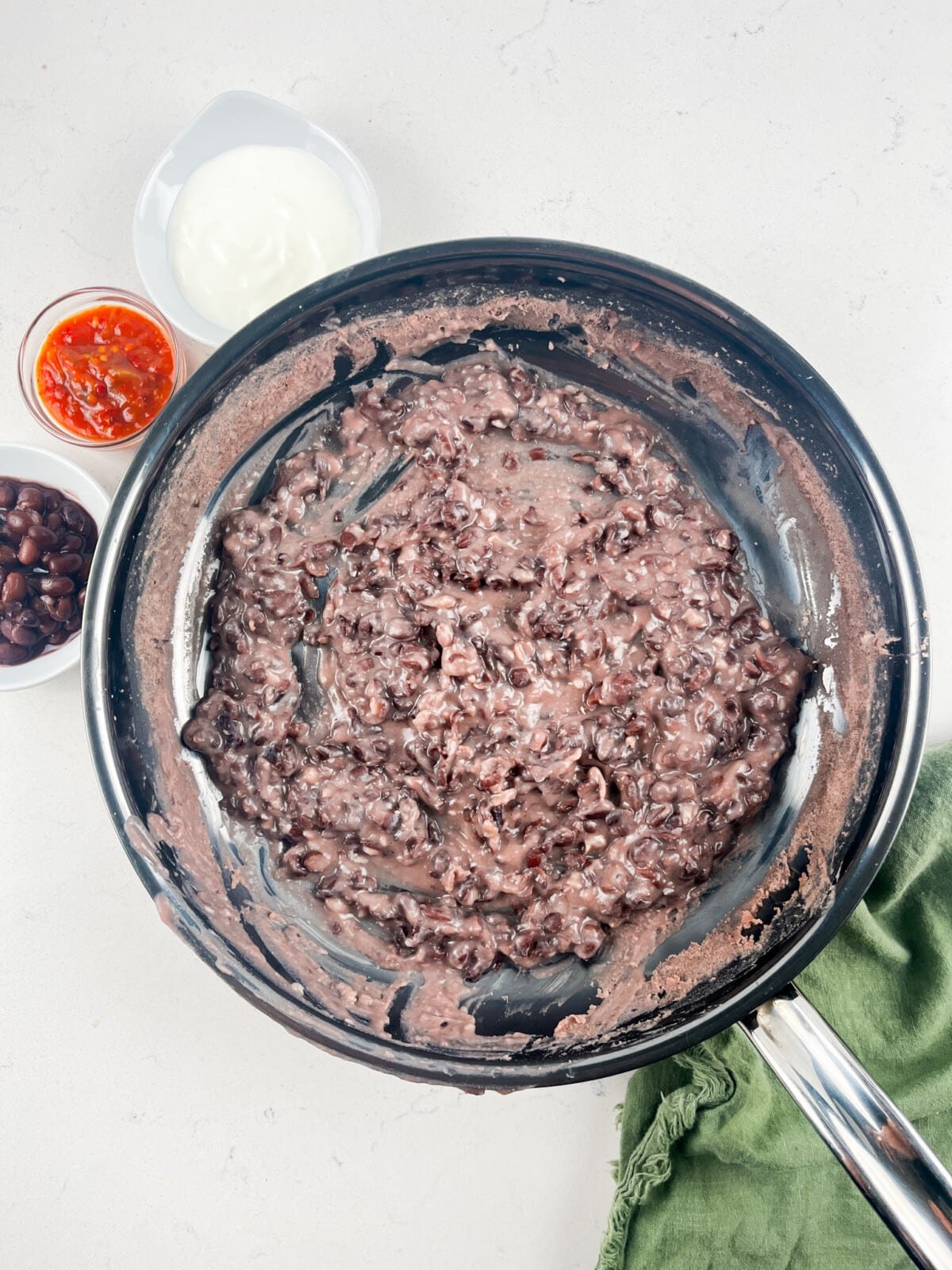 Pan of mashed black beans on a light surface with bowls of sour cream, black beans, and salsa nearby. A green cloth is partially visible.