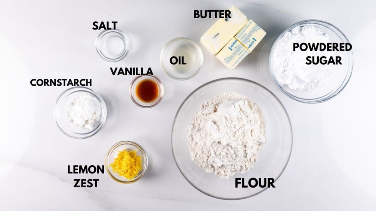Top-down view of labeled baking ingredients on a white surface: small bowls of cornstarch, lemon zest, salt, vanilla, and oil; larger bowls of flour and powdered sugar; and a stick of butter.