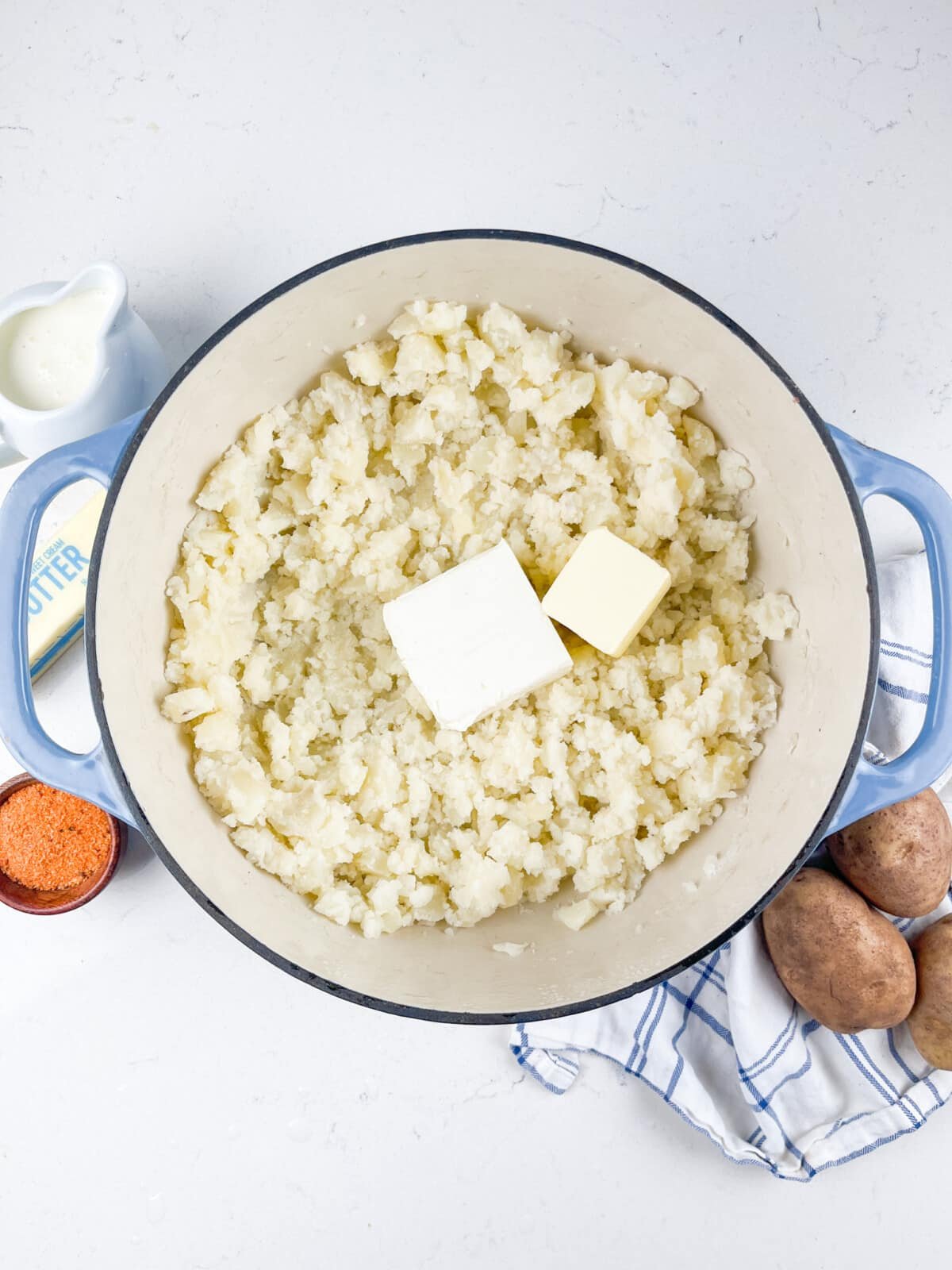A pot of mashed potatoes with two blocks of butter on top. Surrounding the pot are ingredients including cream in a small pitcher, seasoning, and whole potatoes on a cloth. The pot is set on a white surface.
