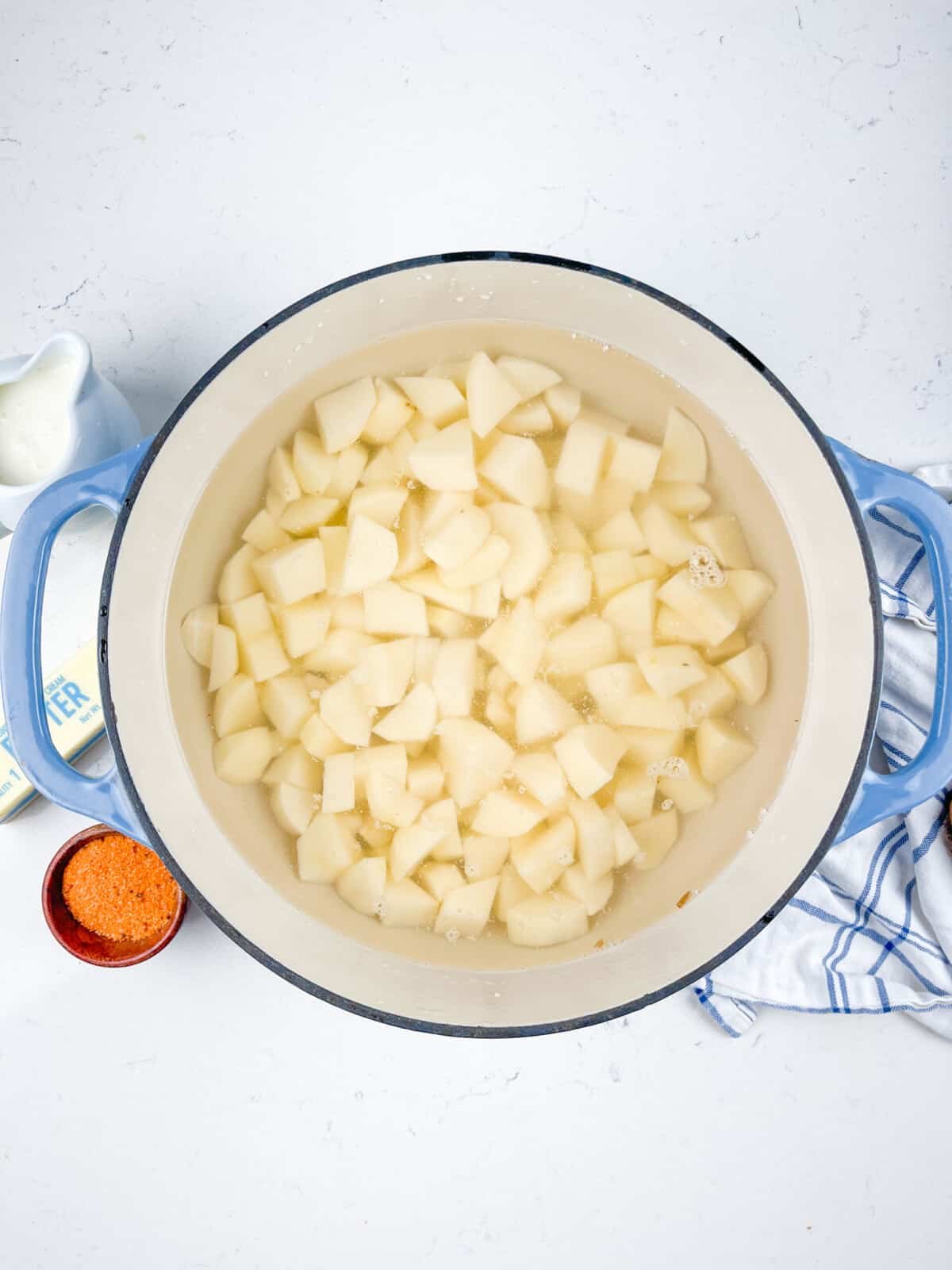A large pot filled with diced potatoes in water, resting on a white countertop. Nearby are a small bowl of orange seasoning, a pitcher of milk, and a striped cloth. The pot has light blue handles.