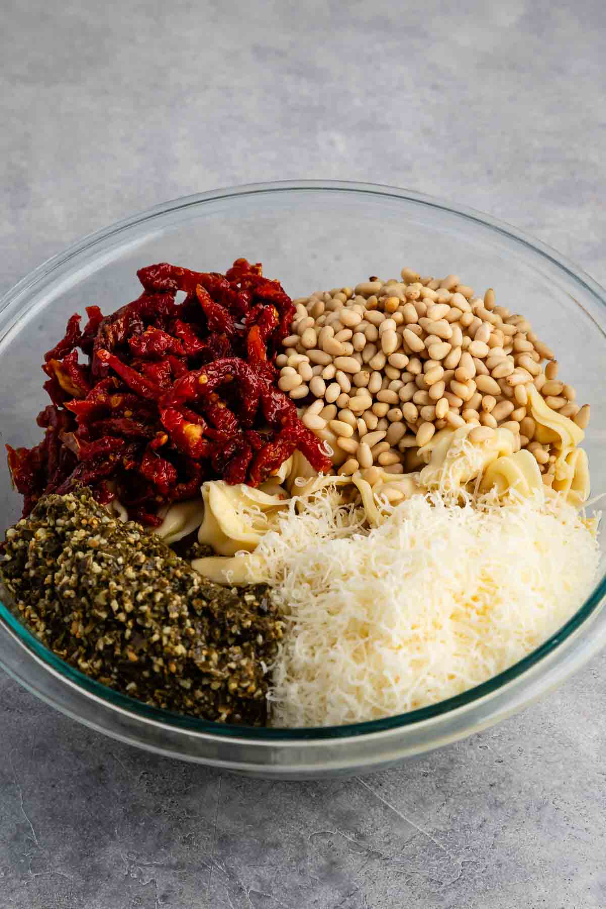 A glass bowl filled with ingredients: sun-dried tomatoes, pine nuts, artichoke hearts, pesto, and grated cheese. The ingredients are arranged in separate sections, creating a colorful and textured display. The background is a gray surface.