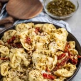 A bowl of tortellini pasta mixed with pesto sauce, sun-dried tomatoes, and pine nuts. The dish is garnished with grated cheese. In the background, there are wooden serving utensils and small bowls containing grated cheese and pesto.