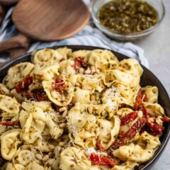 A bowl of tortellini pasta mixed with pesto sauce, sun-dried tomatoes, and pine nuts. The dish is garnished with grated cheese. In the background, there are wooden serving utensils and small bowls containing grated cheese and pesto.