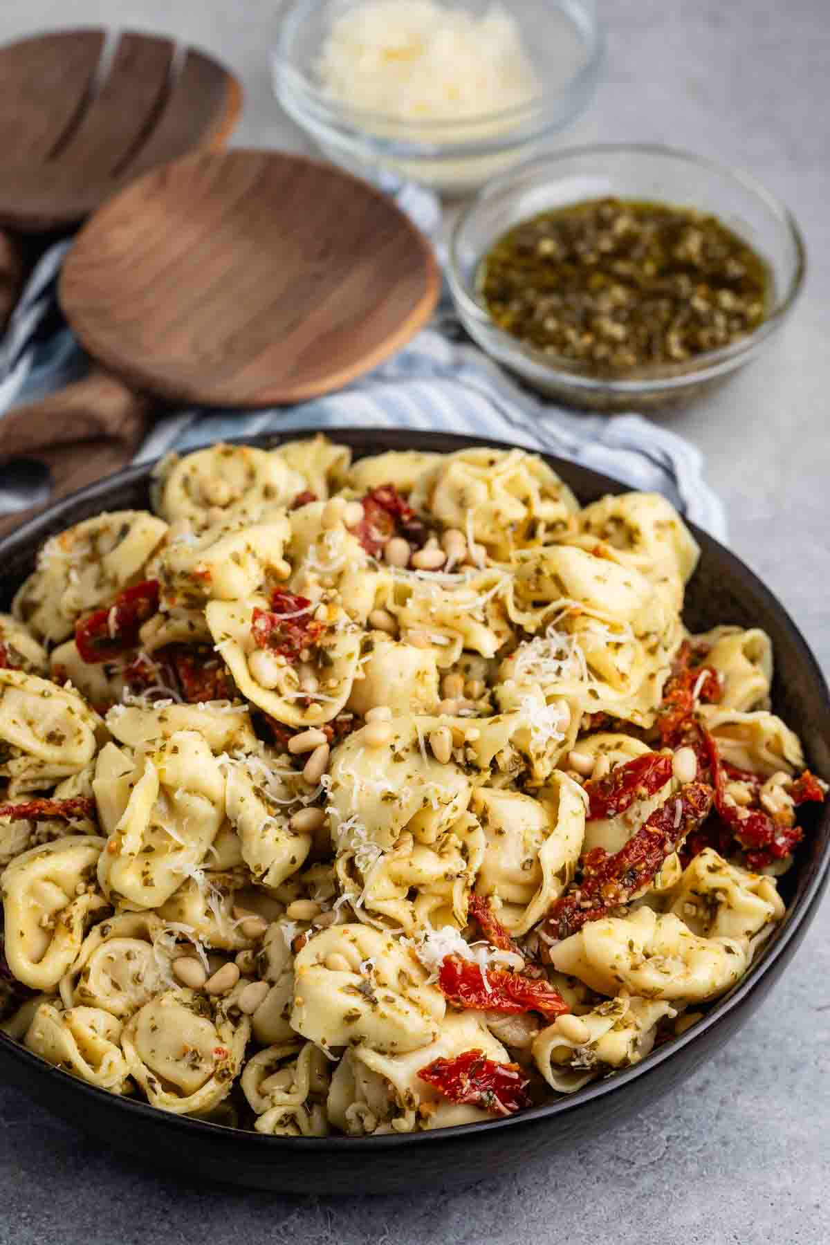 A bowl of tortellini pasta mixed with pesto sauce, sun-dried tomatoes, and pine nuts. The dish is garnished with grated cheese. In the background, there are wooden serving utensils and small bowls containing grated cheese and pesto.