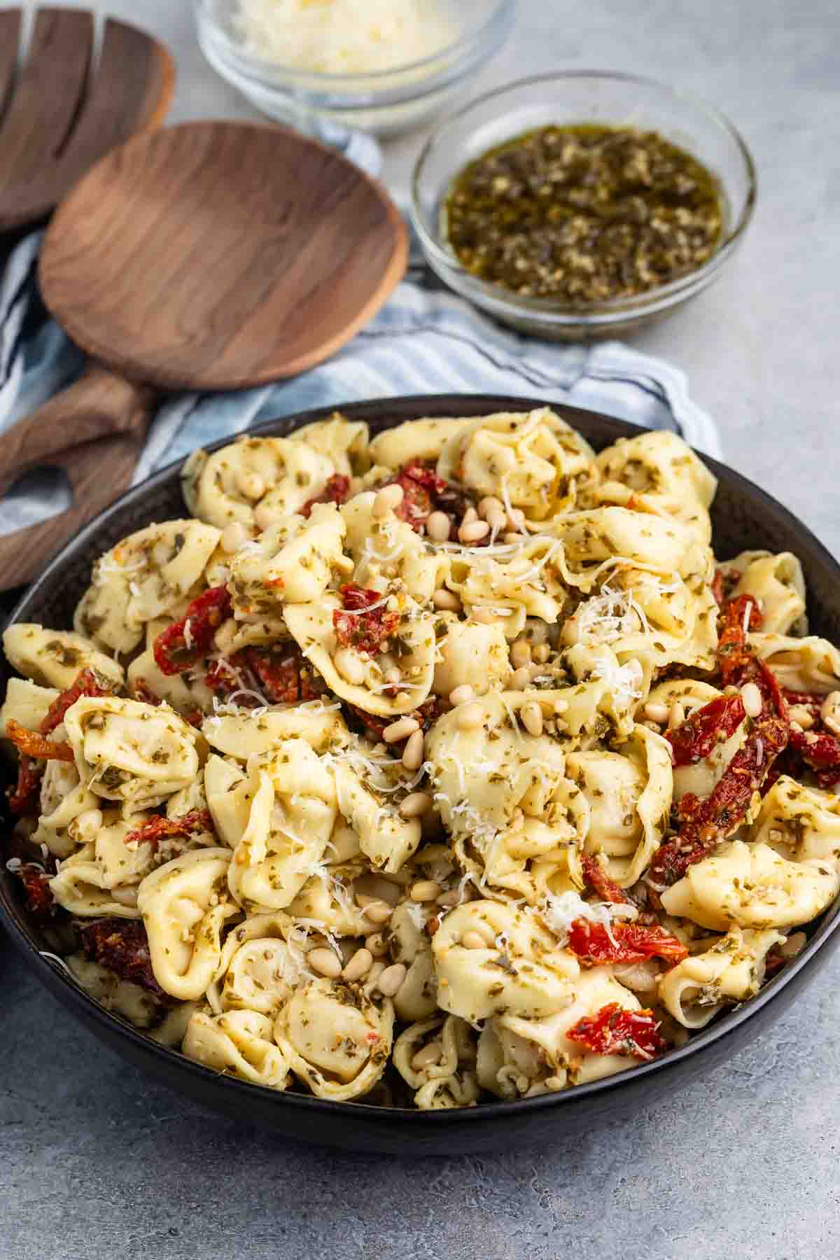 A bowl of tortellini pasta is topped with pesto, sun-dried tomatoes, and pine nuts. In the background, theres a small bowl of extra pesto, a bowl of grated cheese, and wooden serving utensils on a striped cloth.