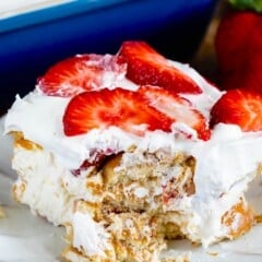 A slice of strawberry shortcake on a white plate, topped with fresh strawberry slices and whipped cream. A large dish with the dessert is visible in the background, along with a whole strawberry on the side.