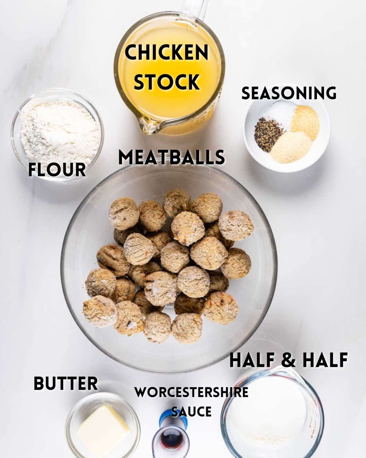 Top-down view of ingredients for a dish: a measuring cup with chicken stock, a bowl of meatballs, small bowls of seasoning and flour, a plate with butter, a glass with half & half, and a bowl labeled Worcestershire sauce on a marble surface.