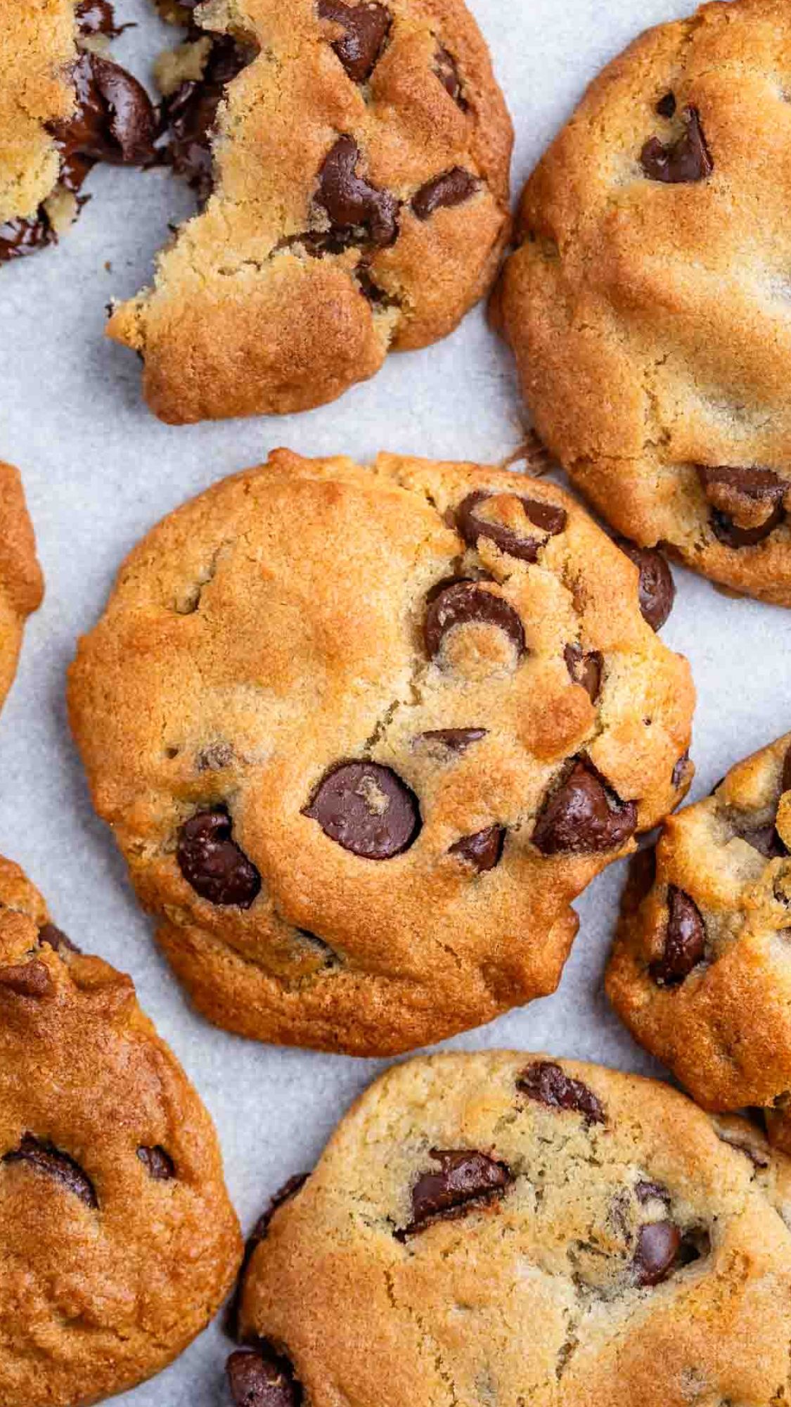 Close-up of several chocolate chip cookies on a light surface. One cookie is broken, revealing its soft interior. The cookies are golden brown with visible chocolate chunks.