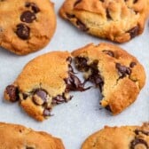 Close-up of chocolate chip cookies on a baking sheet. One cookie is broken in half, showing melted chocolate chips inside. The cookies have a golden-brown crust with visible chocolate chunks.